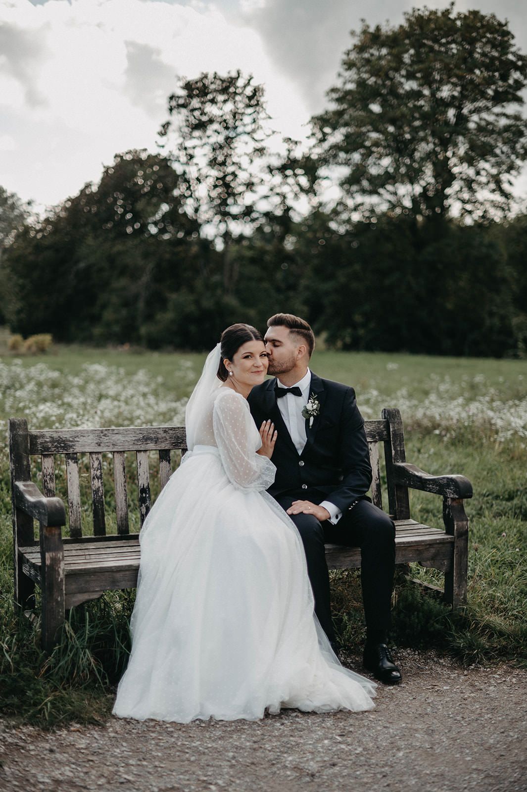 A romantic wedding couple sits together on a rustic wooden bench in a serene outdoor setting with wildflowers.