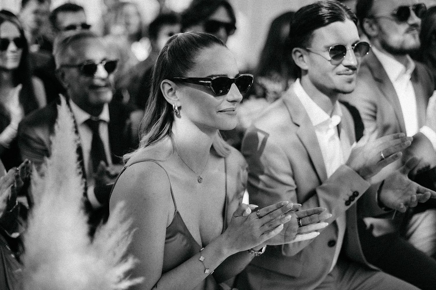 A black and white photo showing people in sunglasses sitting and clapping at an outdoor event.