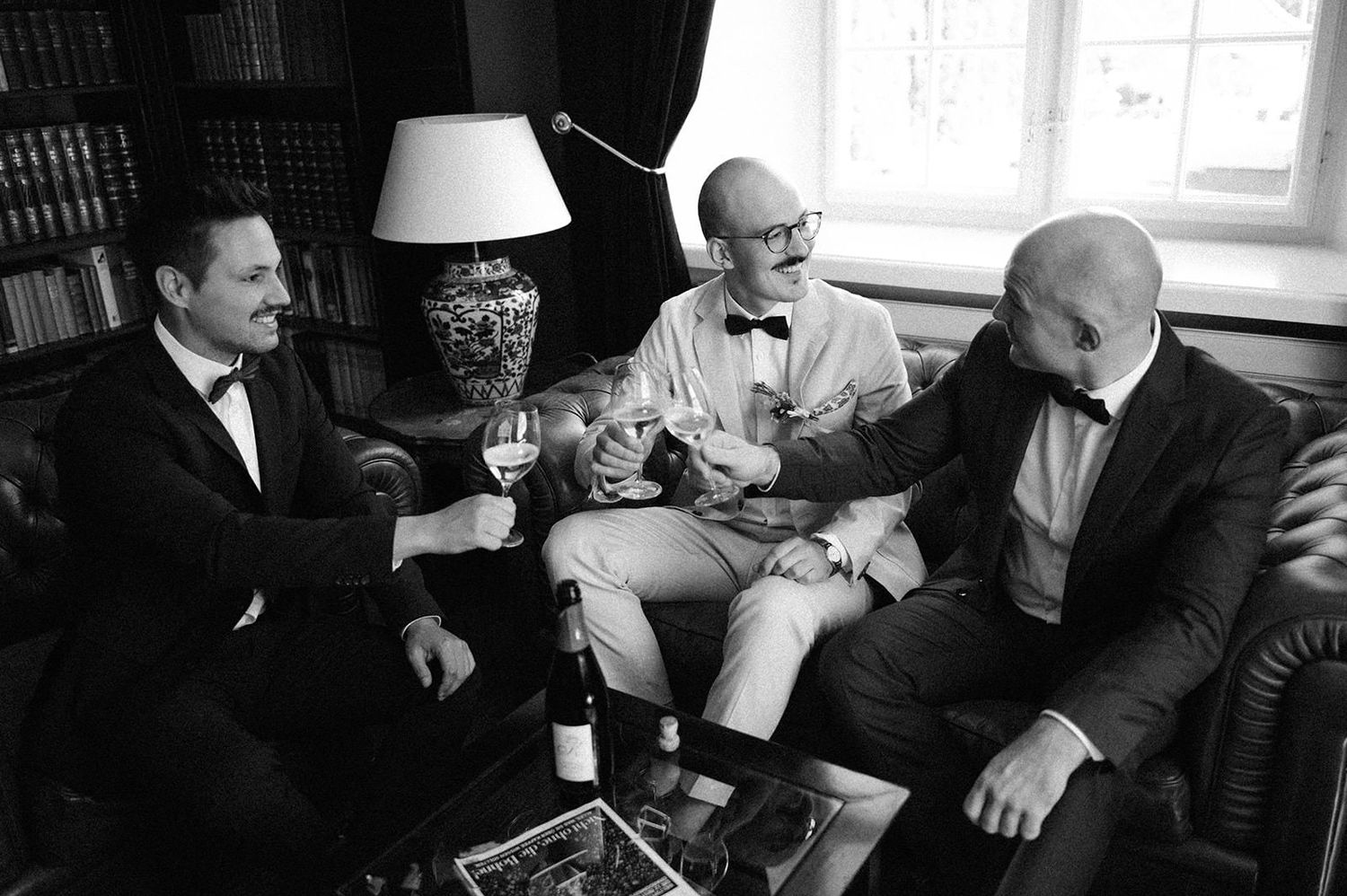 A black and white photo of people gathered in conversation at a social event in a room with windows.