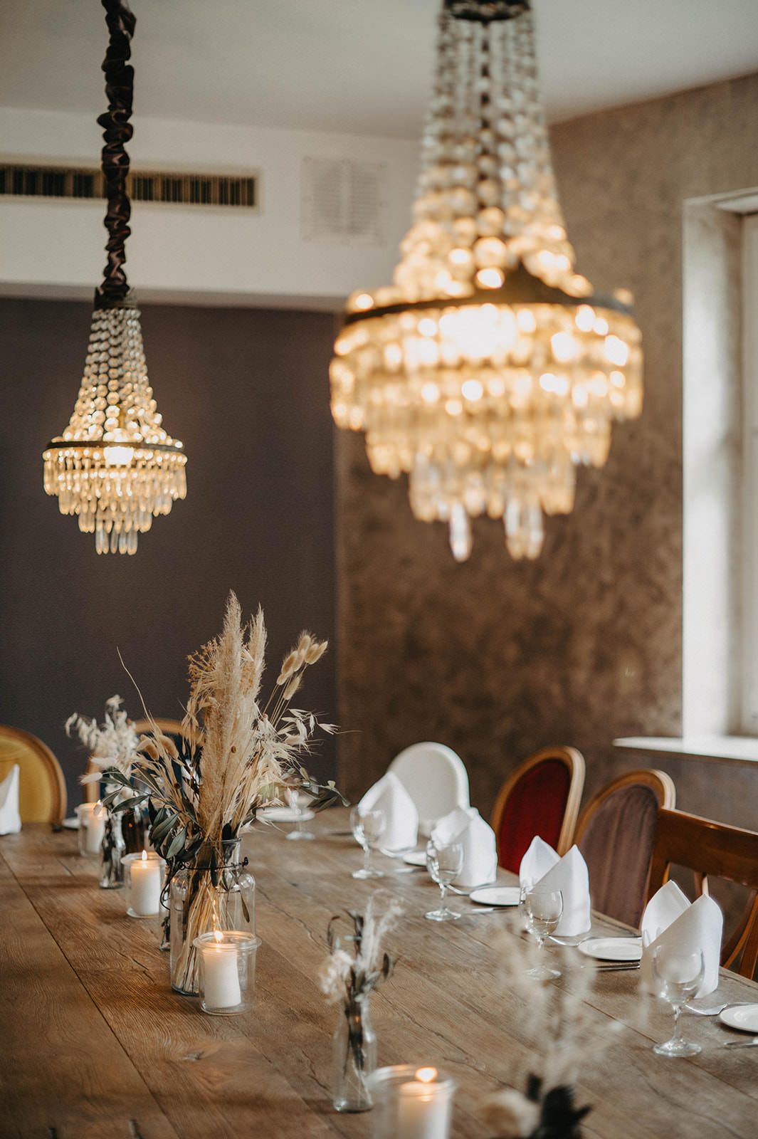 Elegant dining table setup with crystal chandeliers and pampas grass centerpieces in rustic modern setting.