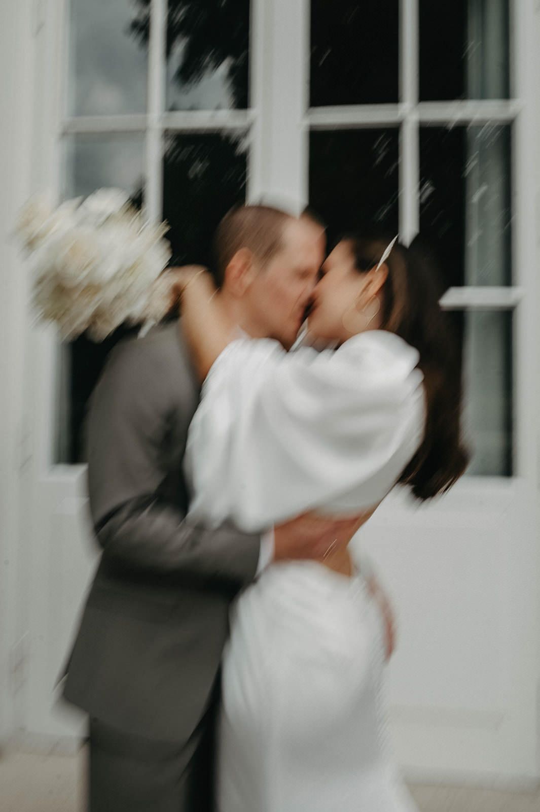 A blurred artistic wedding photo shows a couple sharing a romantic kiss in front of a white window.