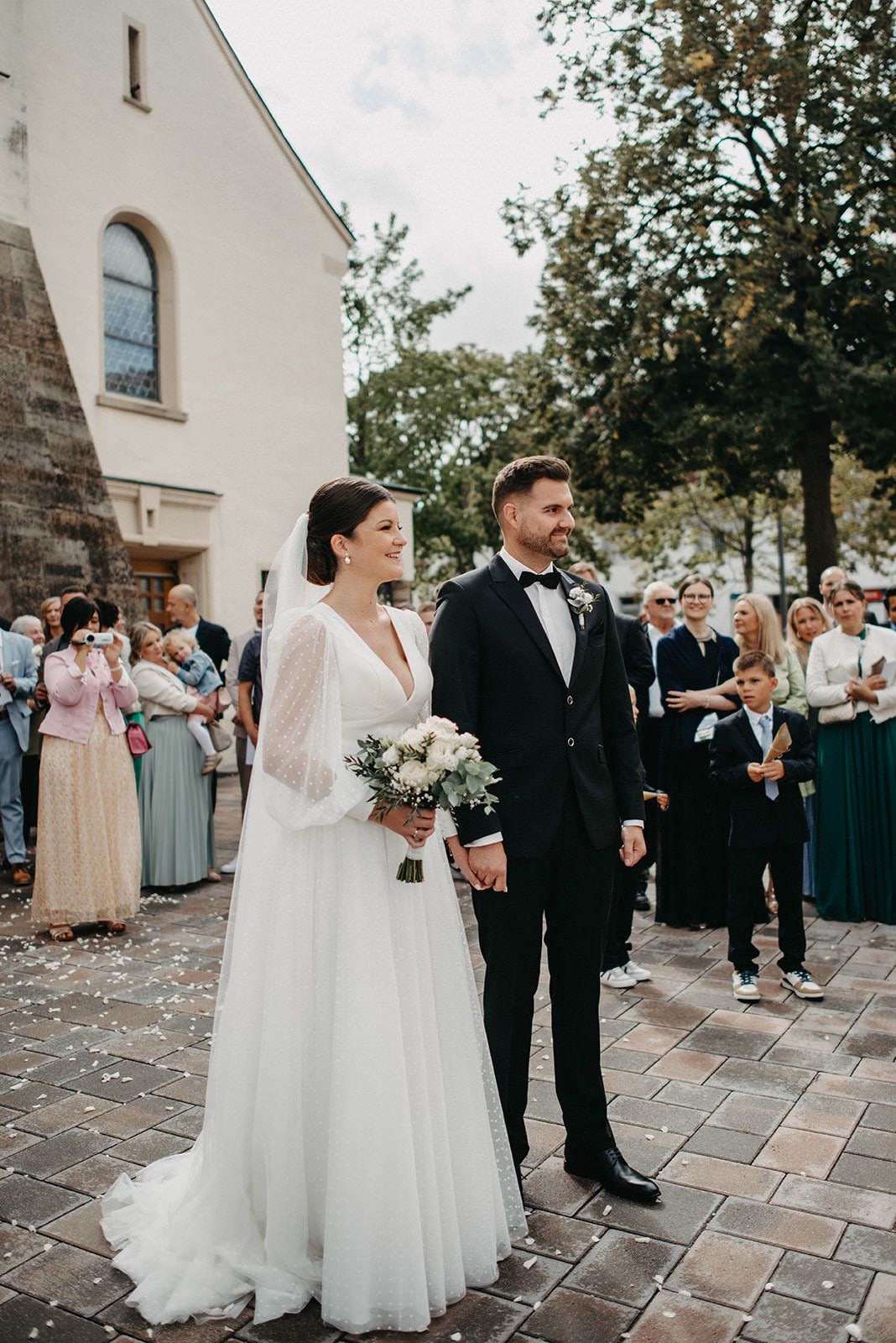 A wedding party stands on stone pavement outside a white church building during a celebration.