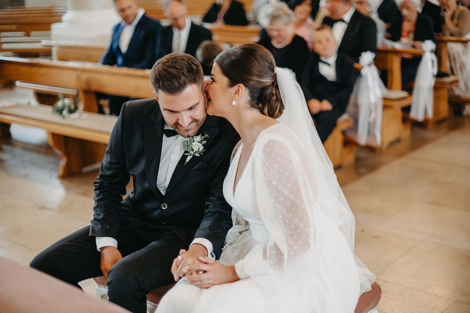 A couple shares an intimate moment during their wedding ceremony while seated in a traditional church pew.