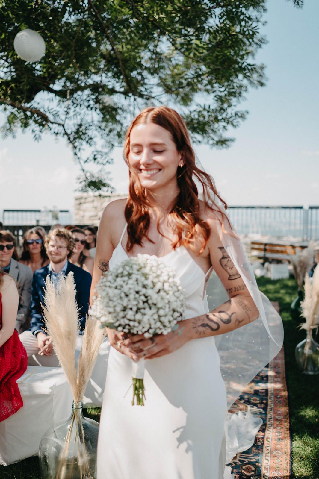 A bride in a white dress holds a bouquet of baby's breath and pampas grass at an outdoor wedding venue.