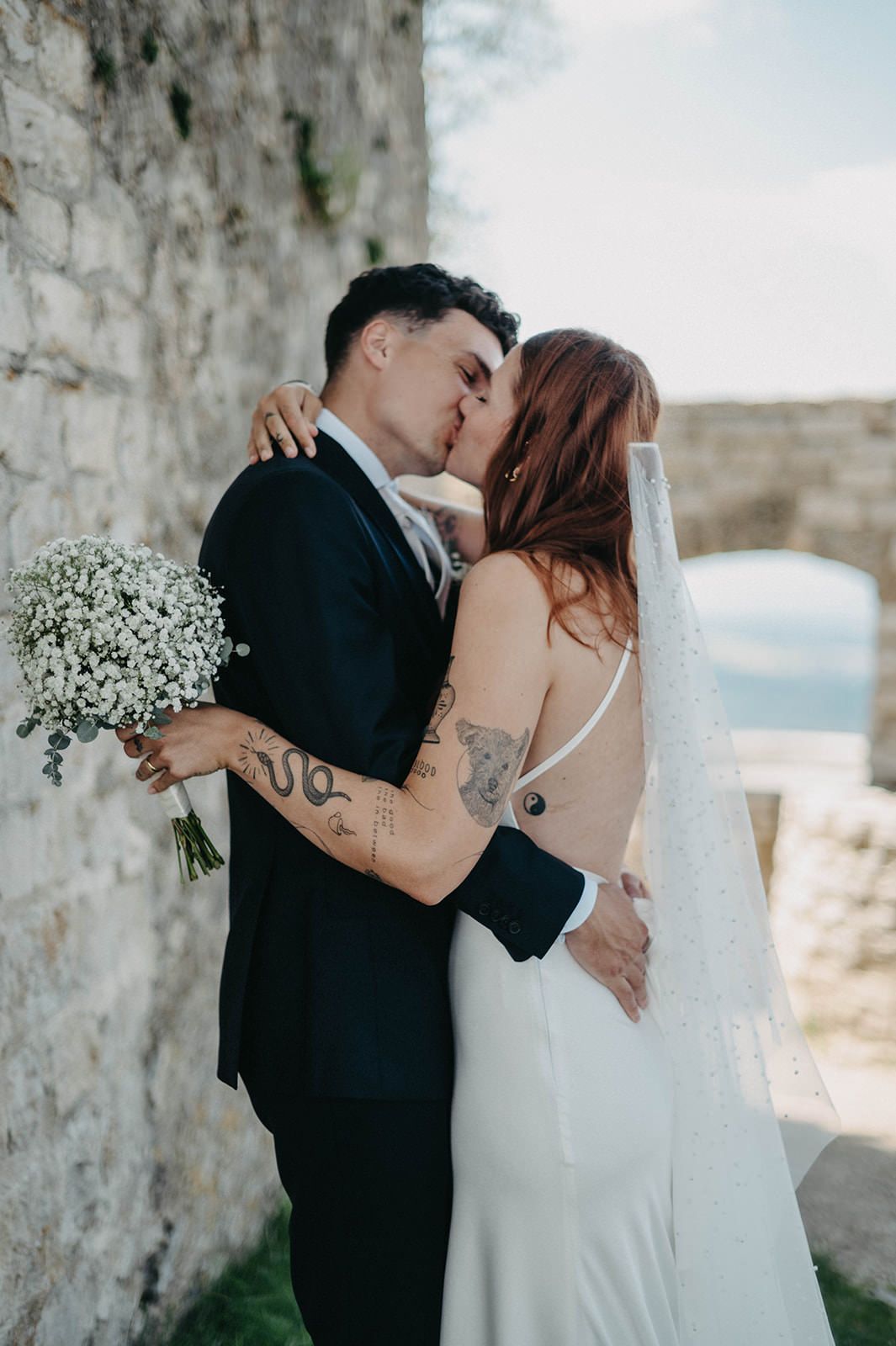 Romantic wedding couple sharing intimate kisses in front of stone wall with baby's breath bouquet and ocean view.