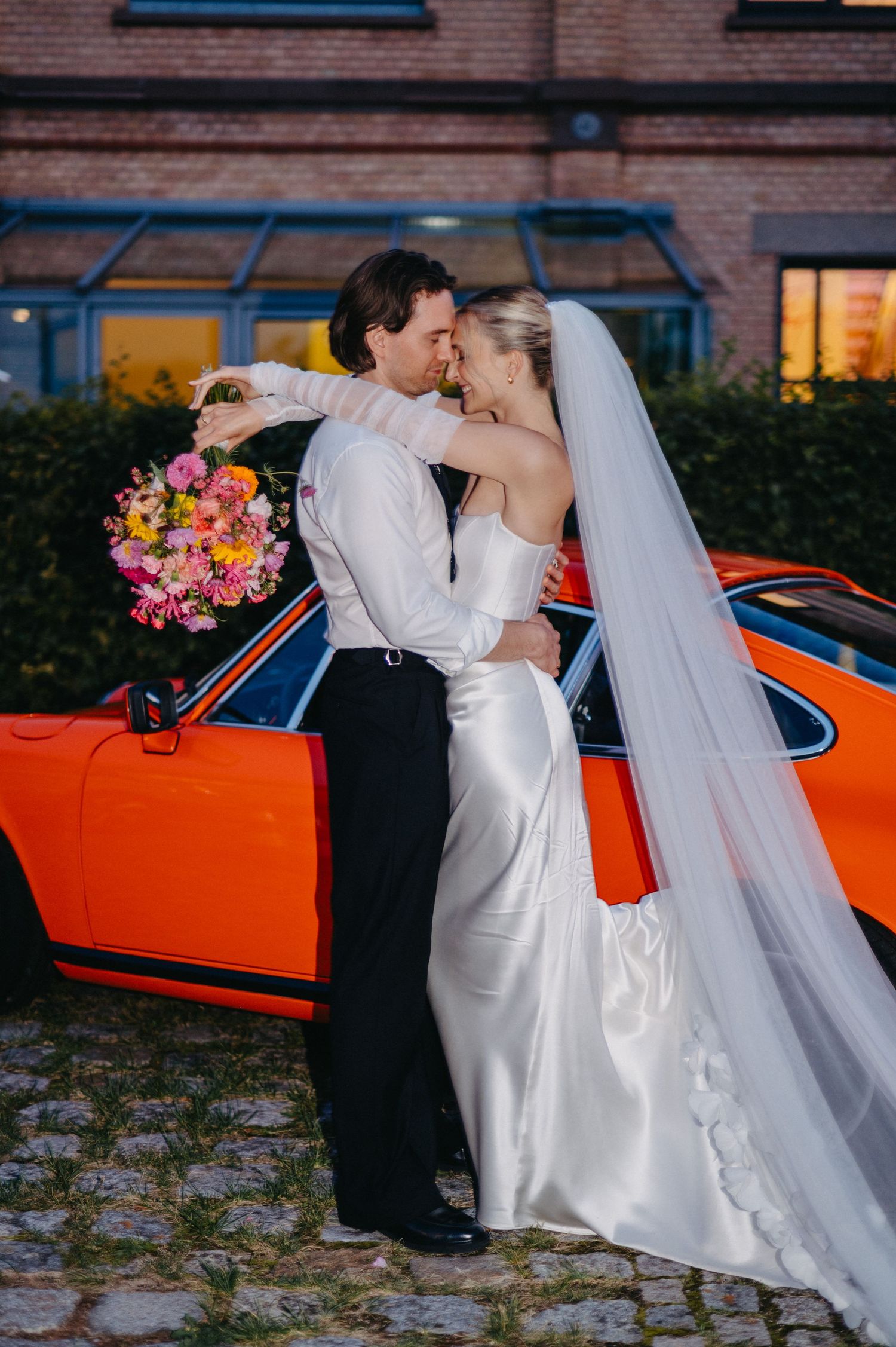 A romantic wedding couple poses with a vibrant orange classic car against a brick building backdrop.