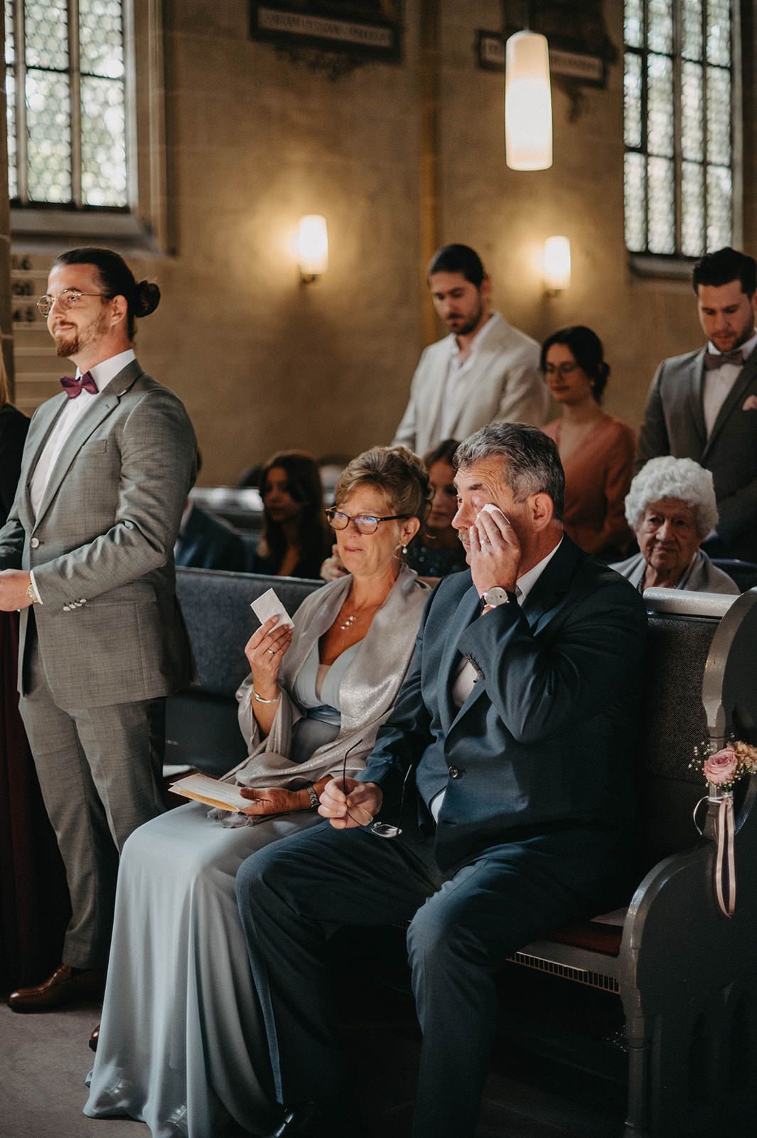Emotional moment during a wedding ceremony in a church with guests seated in pews and soft lighting from wall sconces.