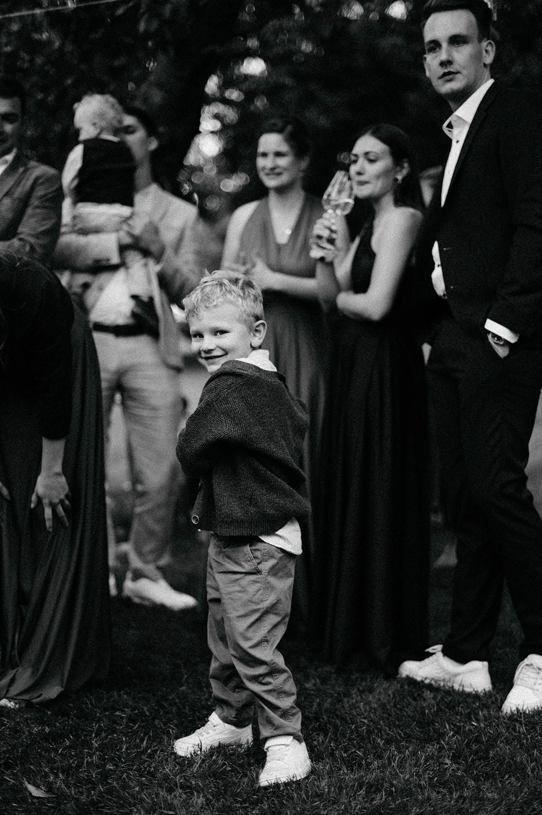 A black and white photograph shows a child in white shoes standing among adults at a formal outdoor gathering.
