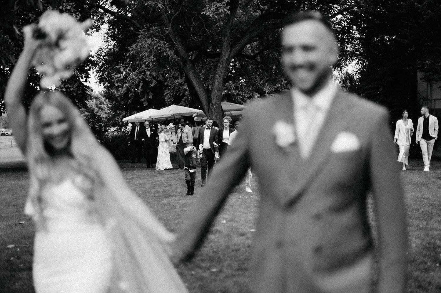 Black and white wedding photo showing a joyful couple holding hands and walking together in a park setting.