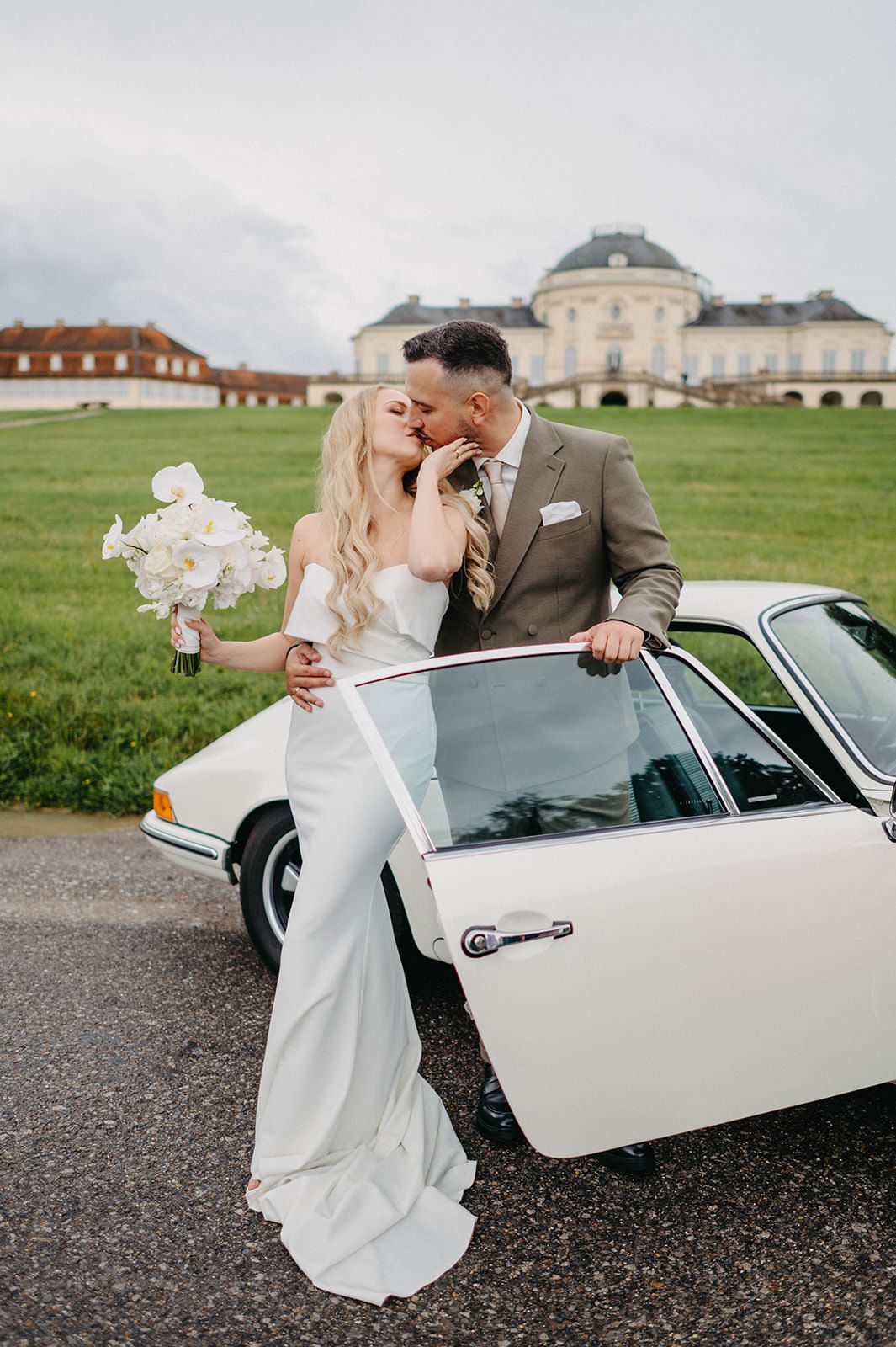 Romantic wedding couple poses with vintage white car outside historic palace grounds.