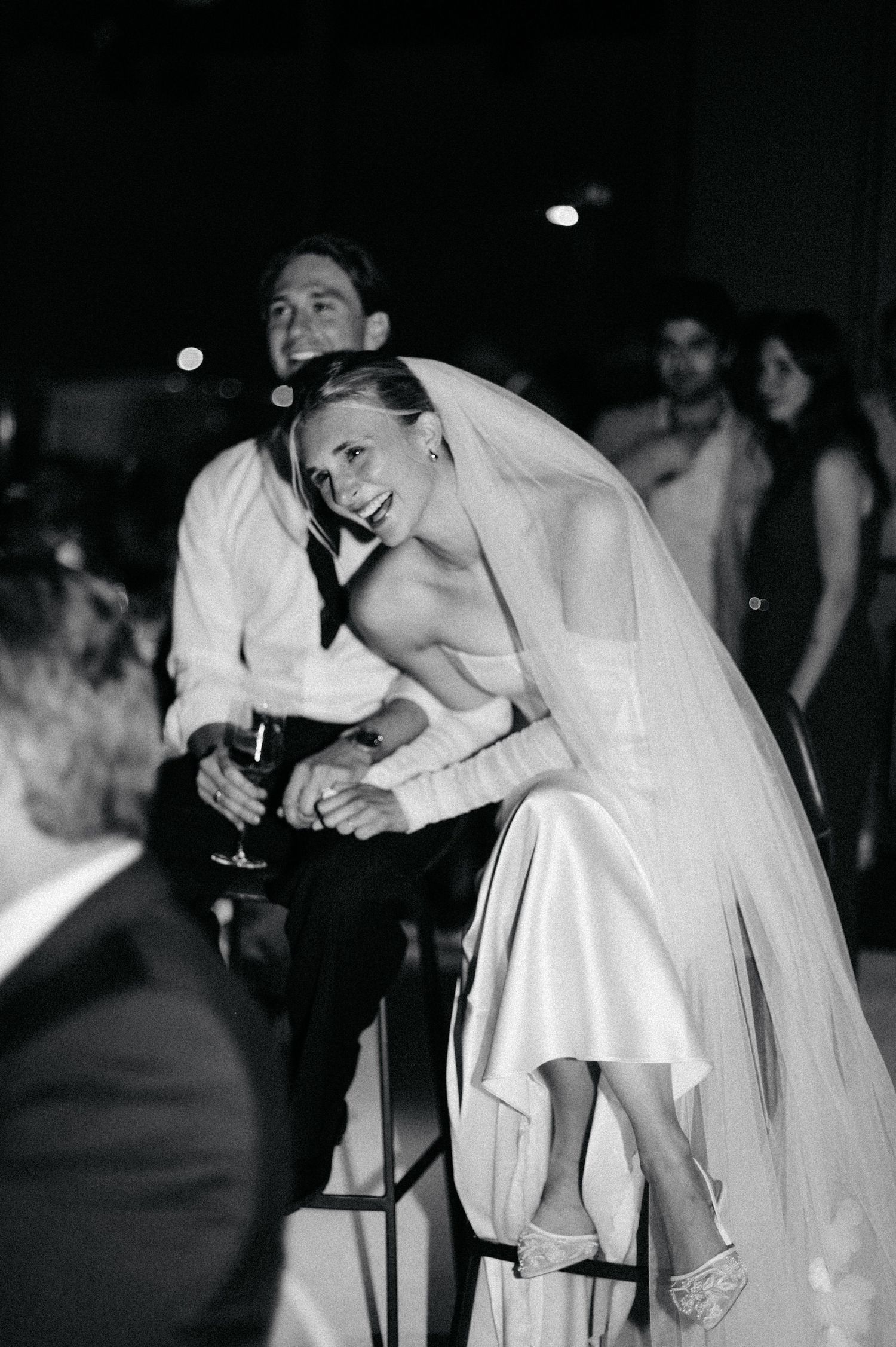 A bride in an elegant white wedding dress leans forward joyfully during a wedding reception celebration.