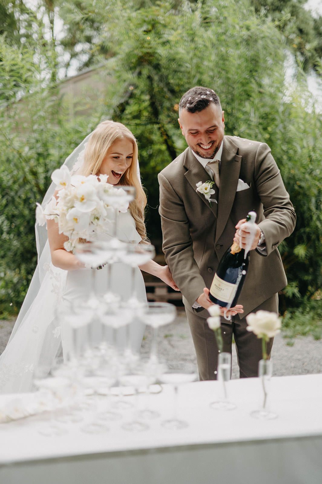 Newlyweds pour champagne into a tower of glasses at an outdoor wedding celebration.