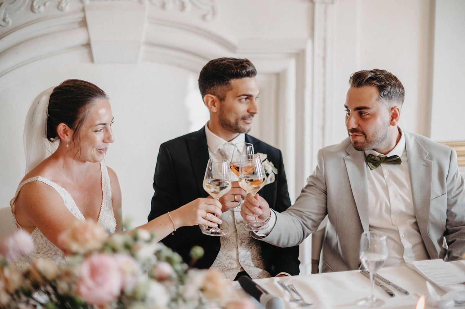 Wedding party members share champagne toasts in an elegant white room with ornate molding and decor.