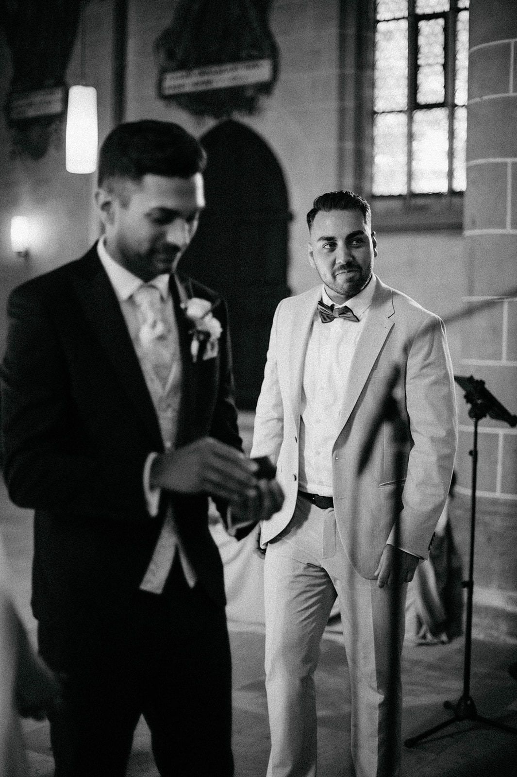 Two men in formal attire stand together at a church wedding, one wearing black and one in light colored suit.