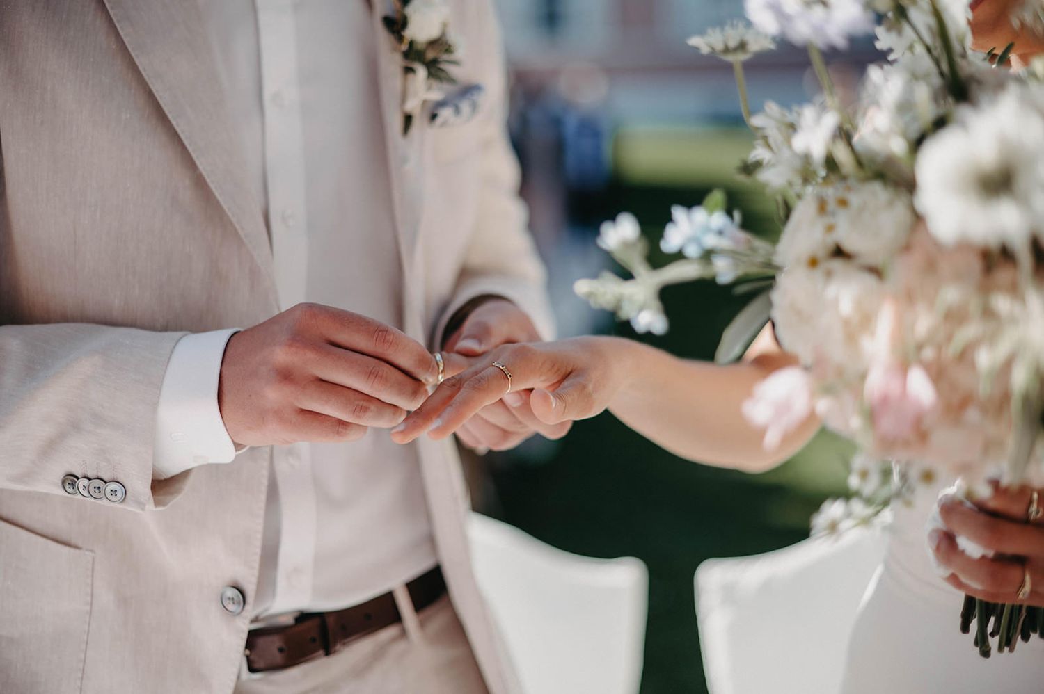 Close-up of hands exchanging wedding rings during an outdoor ceremony with white floral decorations.