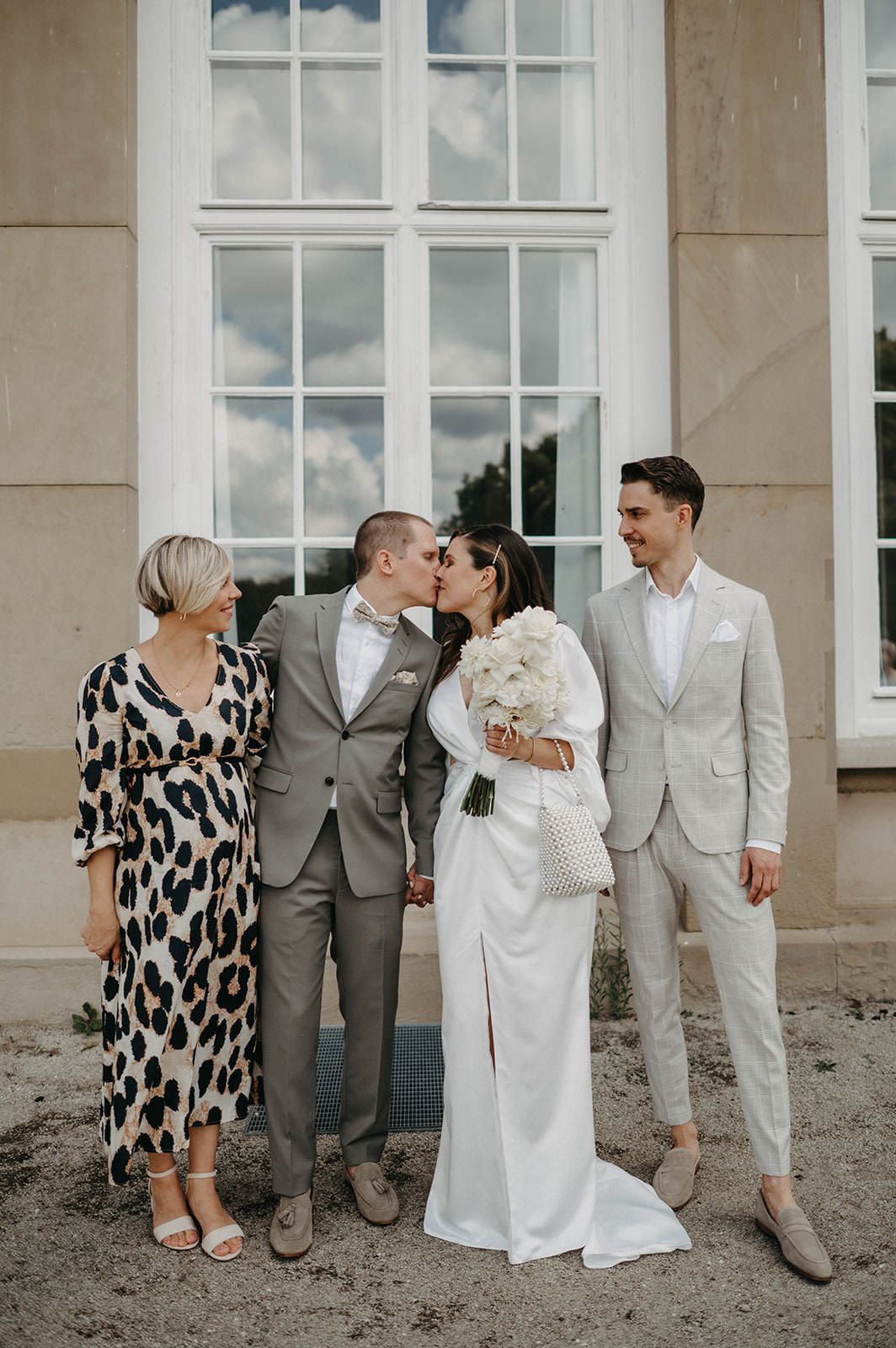 Wedding party in white and grey attire poses together against stone building exterior.