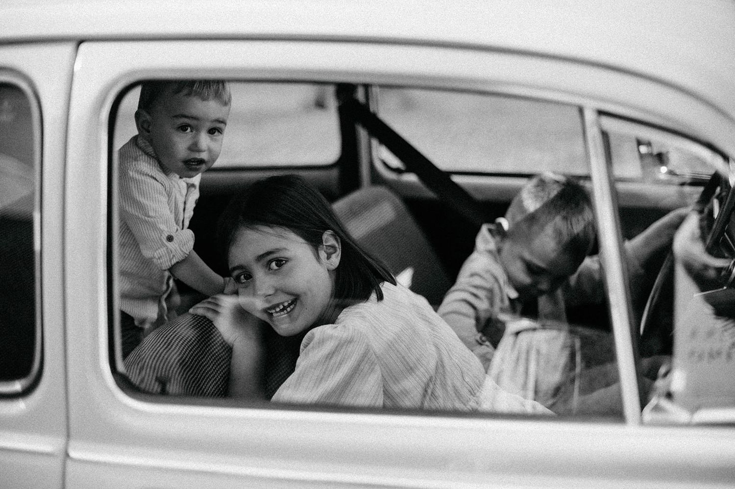 Black and white photo showing a classic car with people inside celebrating at a wedding event.
