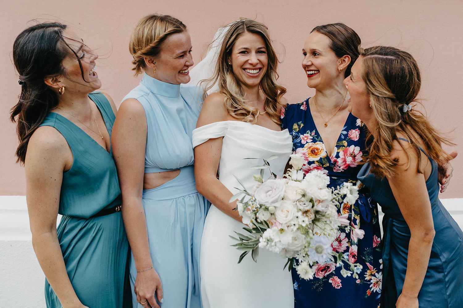 Bridesmaids in various shades of blue dresses share a joyful moment while holding a white floral bouquet.