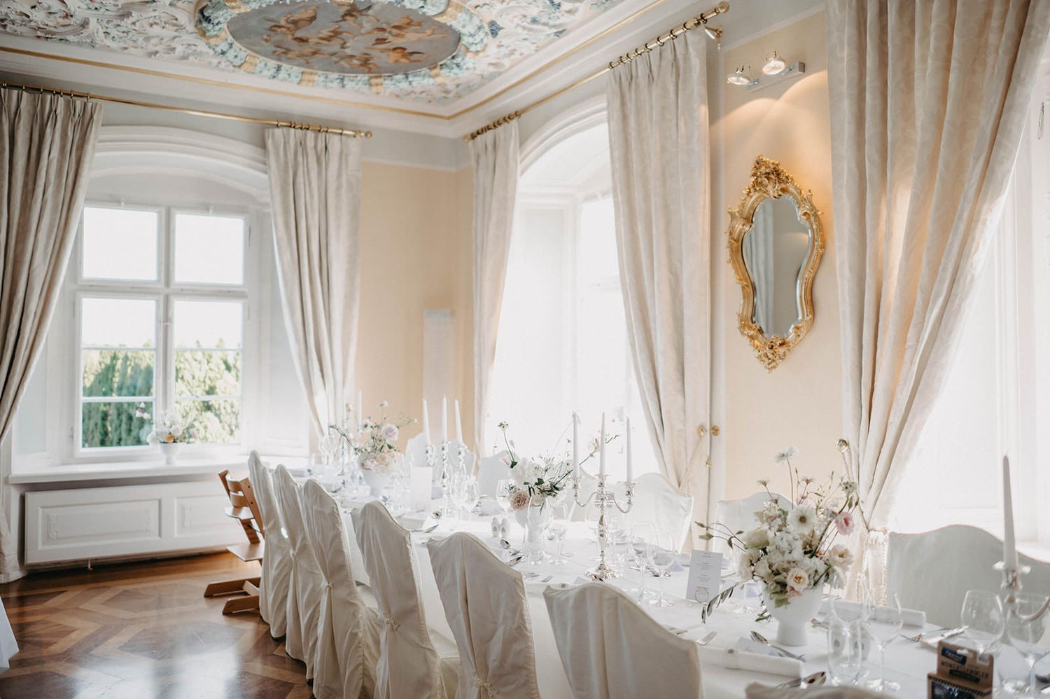 Elegant white wedding reception venue with ornate ceiling details, crystal chandeliers, and floor-to-ceiling windows.