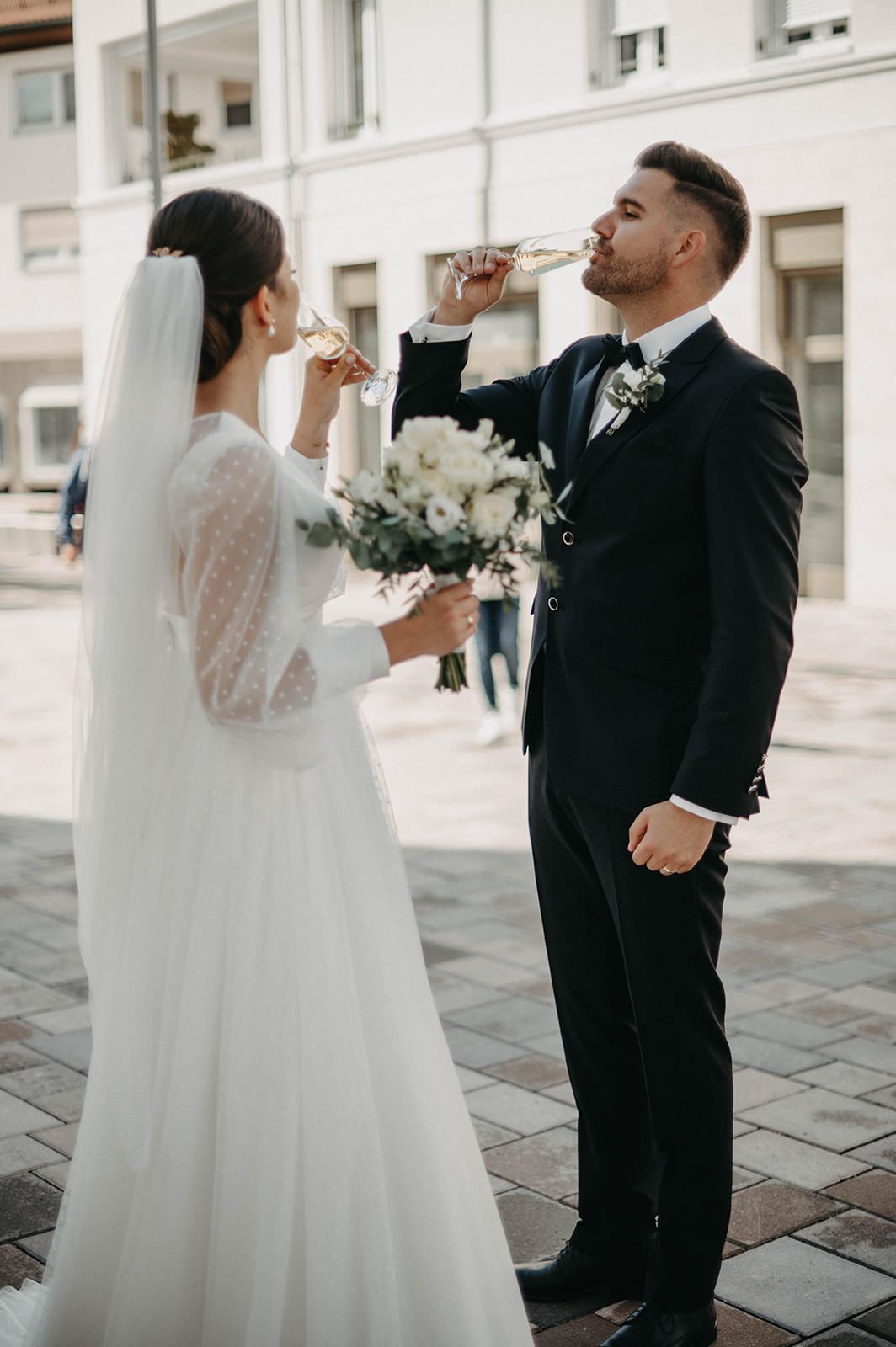 A bride and groom share a drink together on a stone patio during their wedding celebration.