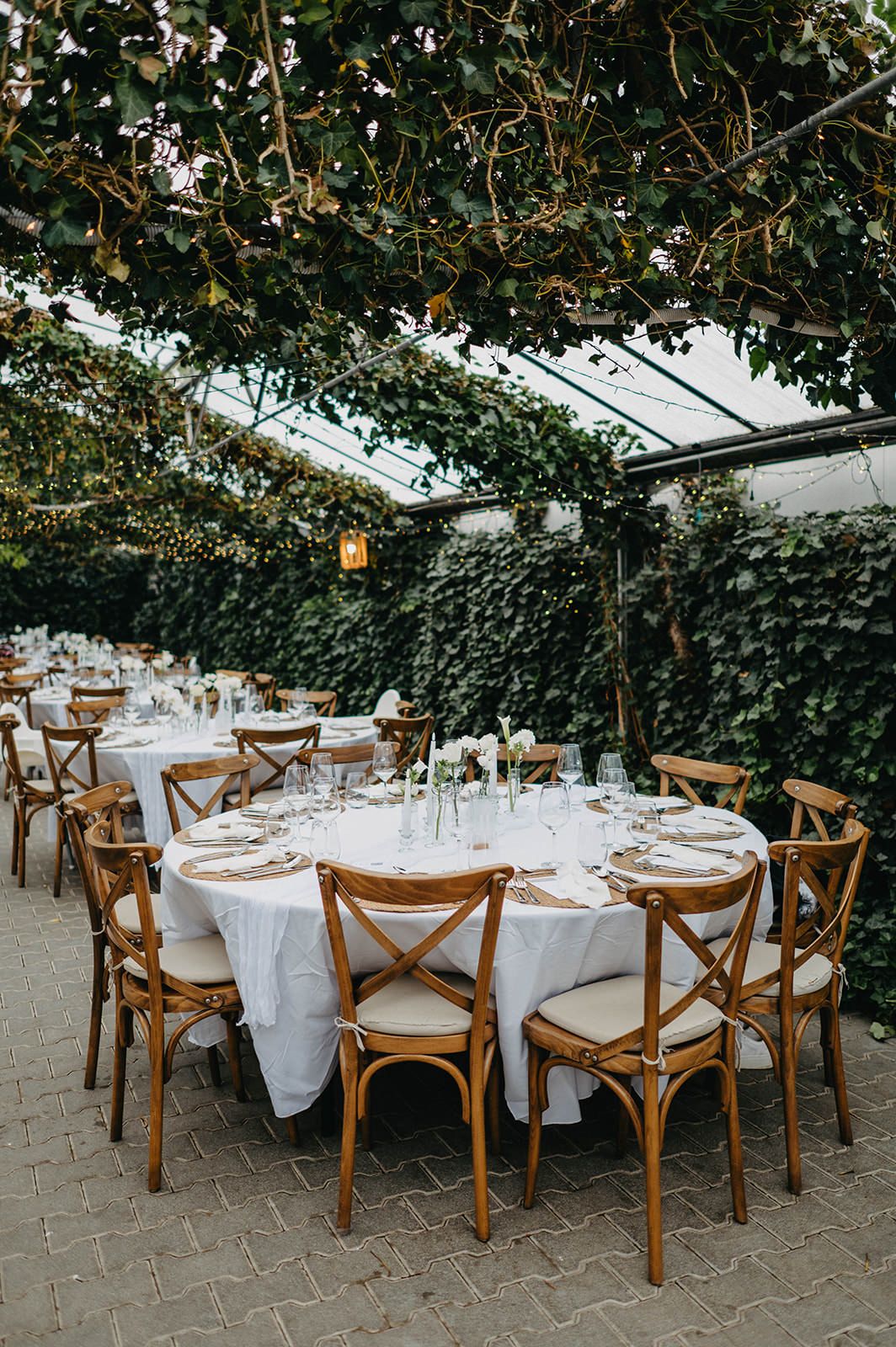 Elegant wedding reception tables set with white linens and wooden cross-back chairs under a lush greenery-covered ceiling.