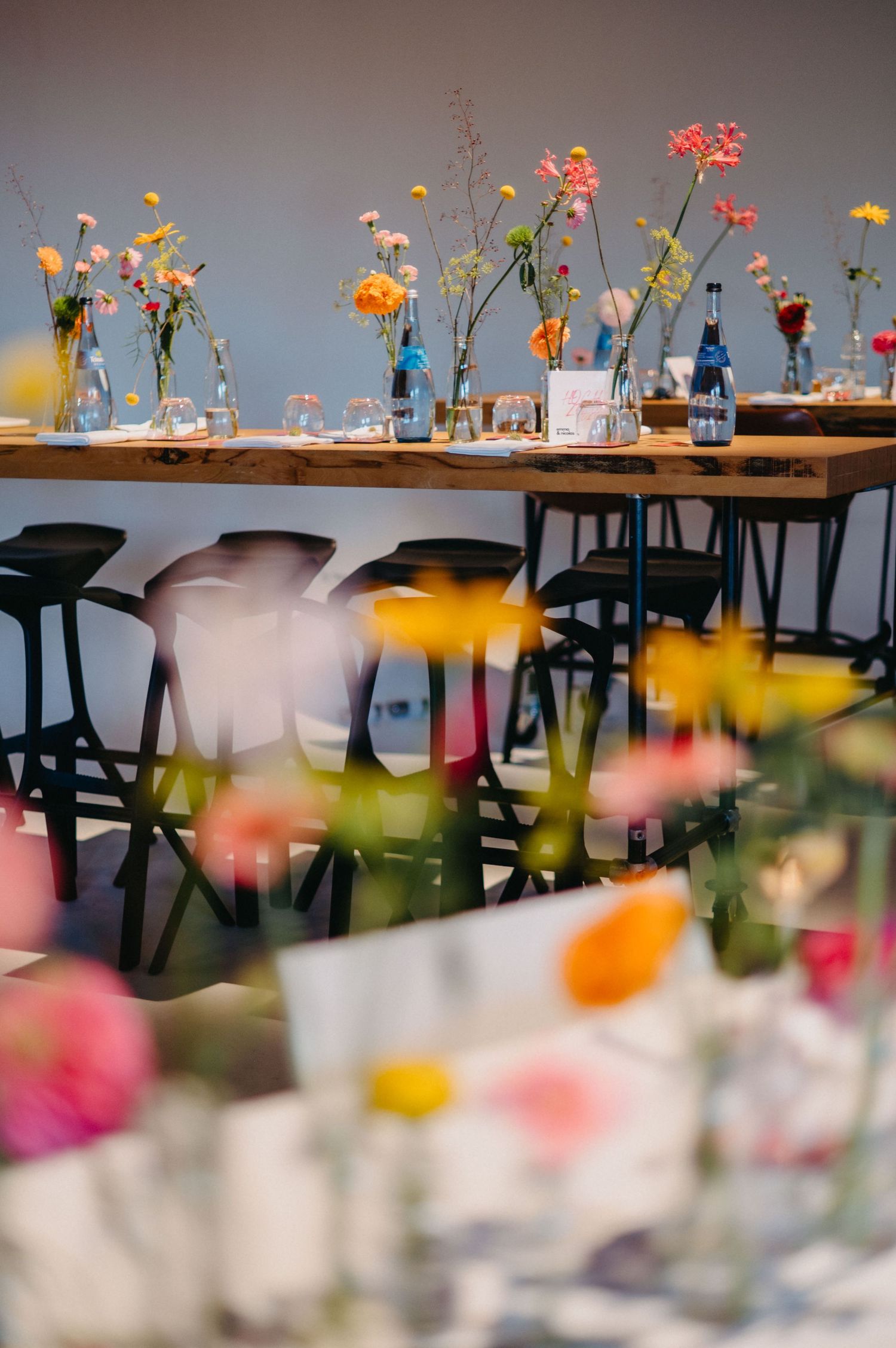Colorful wildflowers in glass vases line a long wooden table with black chairs creating an elegant dining setup.