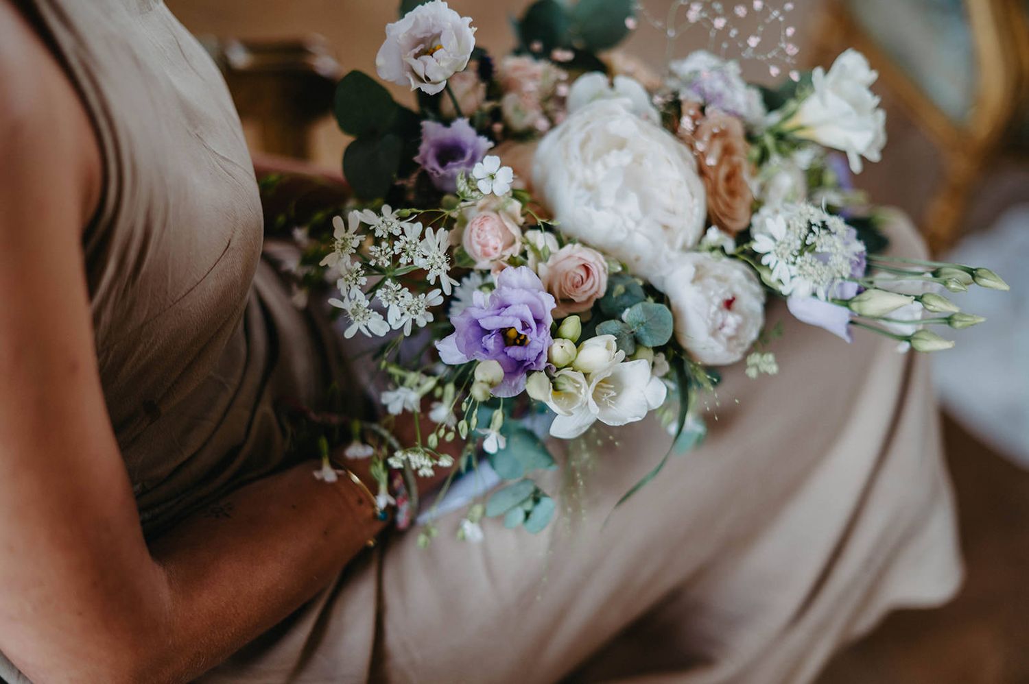 Pastel bouquet with white peonies, purple flowers and baby's breath held against a taupe bridesmaid dress.