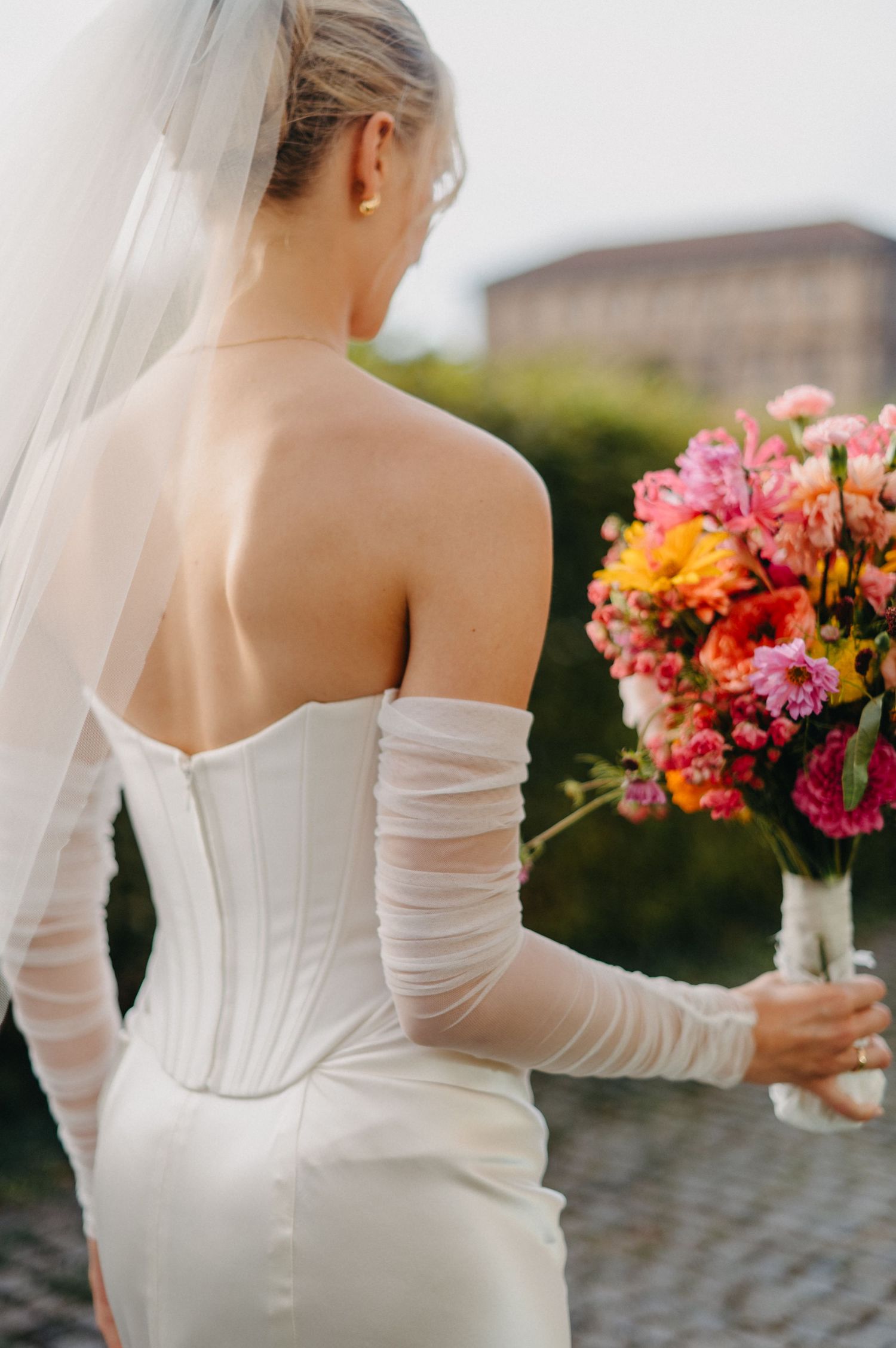 Back view of bride in elegant white strapless gown holding vibrant pink and orange floral bouquet at outdoor wedding.