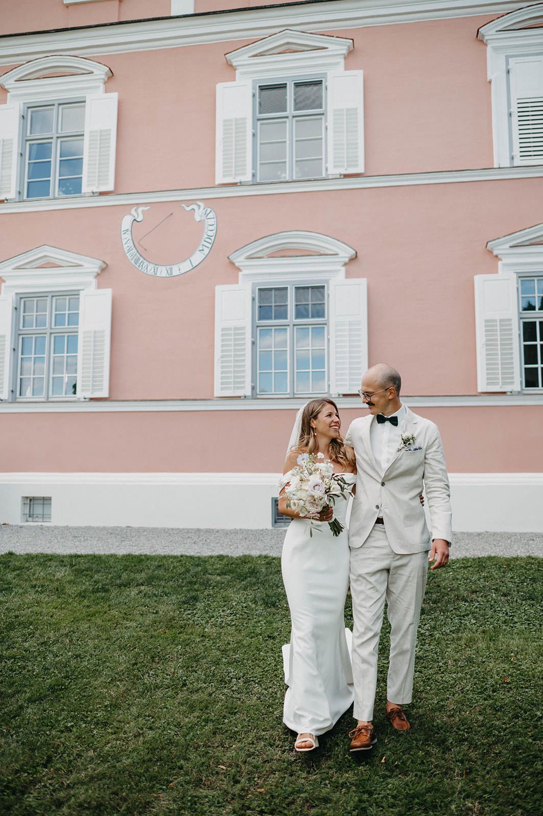 A couple in white wedding attire walking along a pink building exterior with classic architectural details.