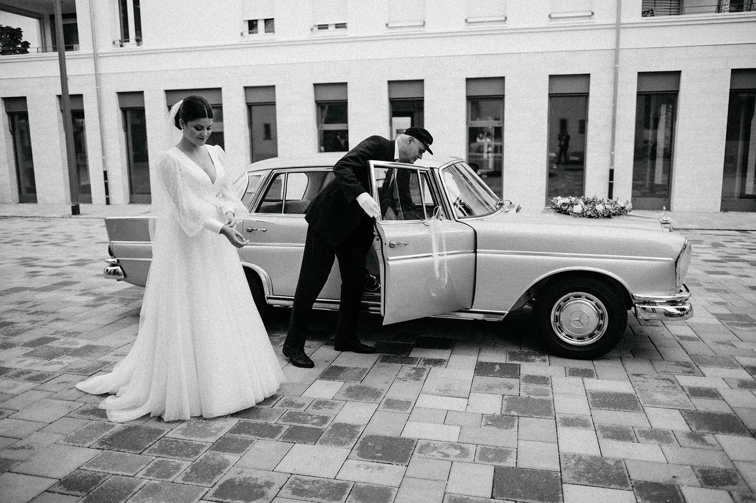 A black and white wedding photograph showing a couple walking together through an elegant courtyard with classic architecture.