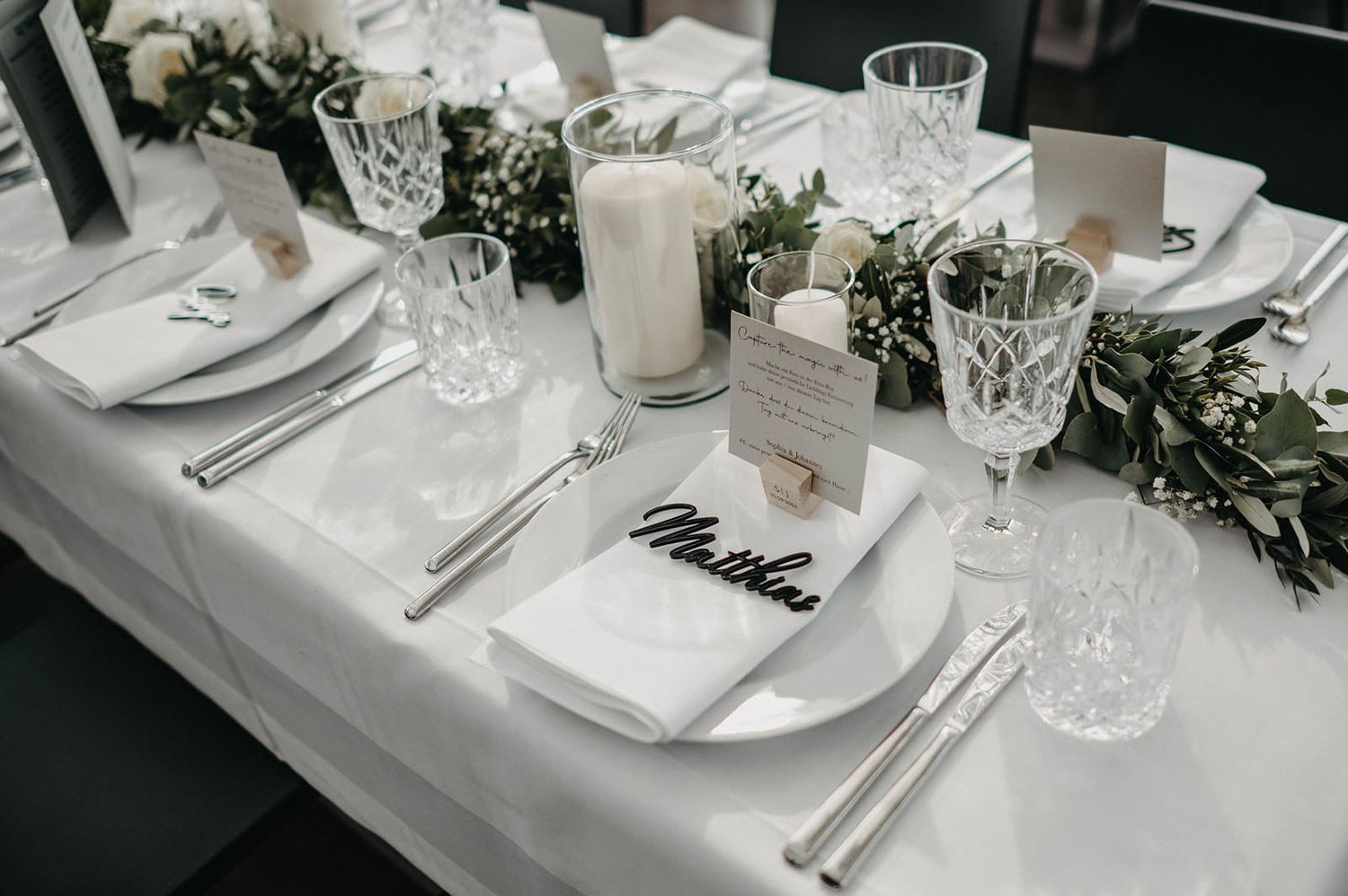 Elegant wedding table settings with white linens, crystal glassware, and delicate greenery decorations at a formal reception.