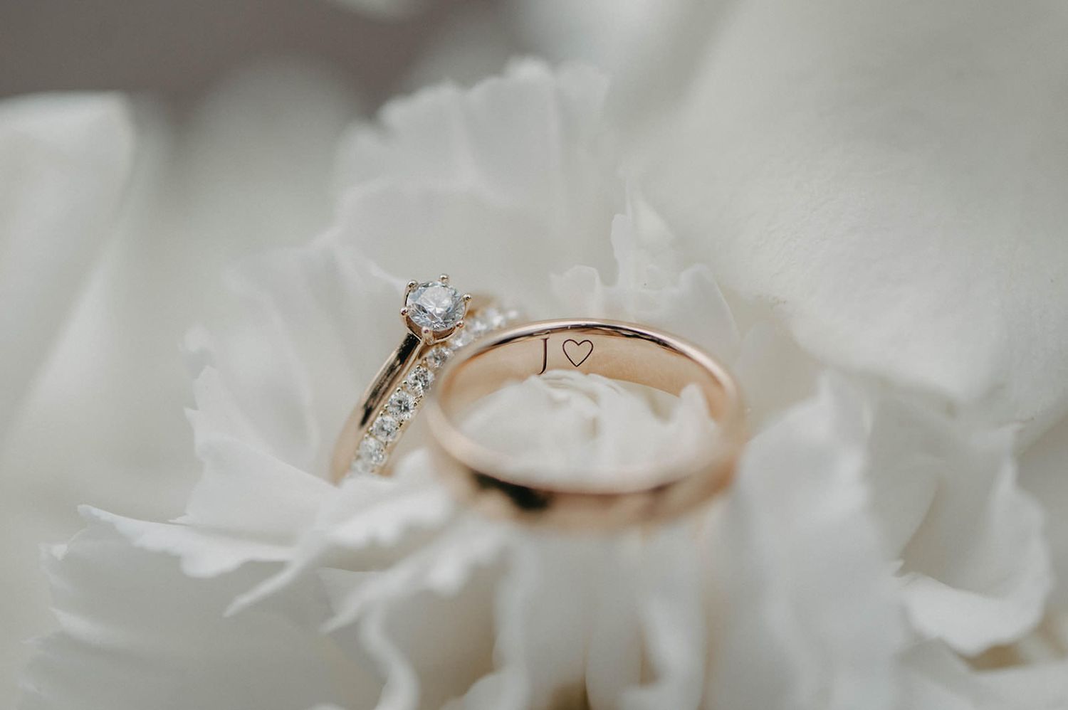 Rose gold engagement ring with diamond solitaire rests delicately on white flower petals in soft focus.
