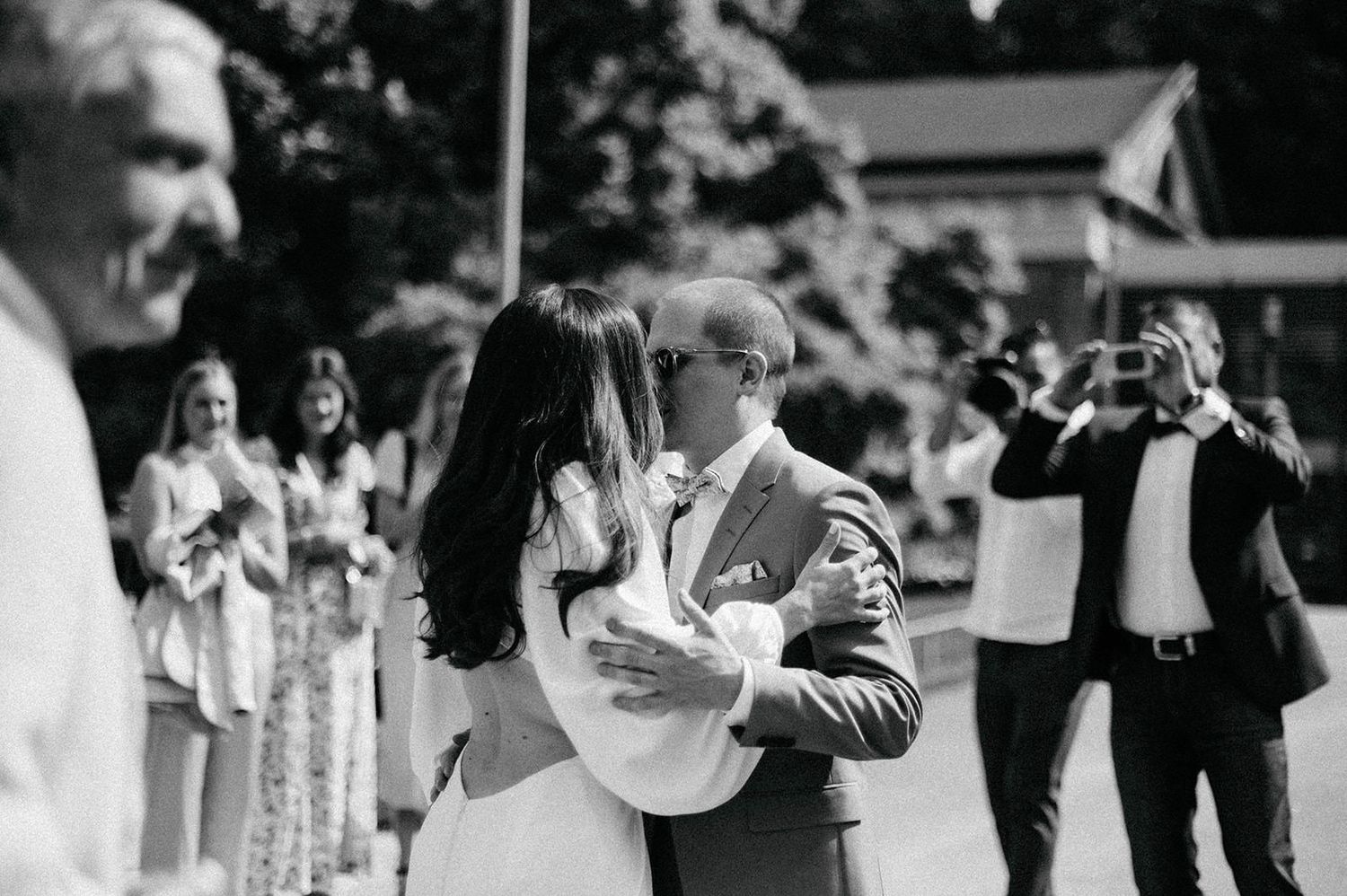A black and white wedding photograph showing a couple dancing while guests watch in the background at an outdoor celebration.