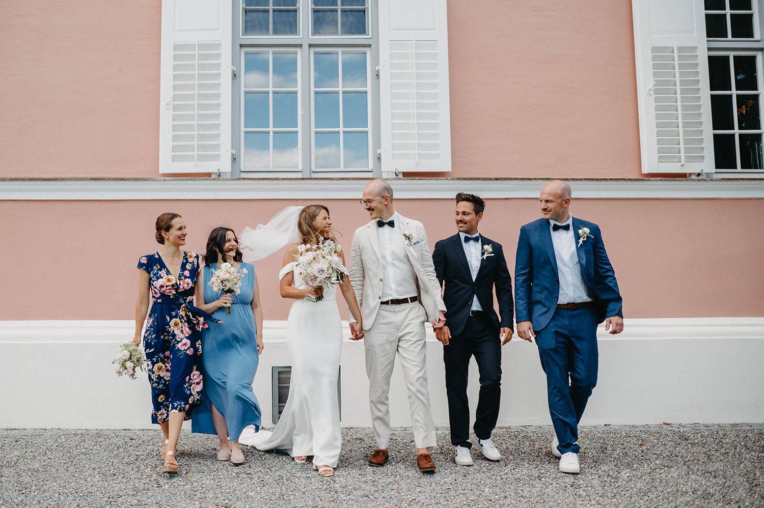 Wedding party walks along pink building exterior while holding hands and laughing together.