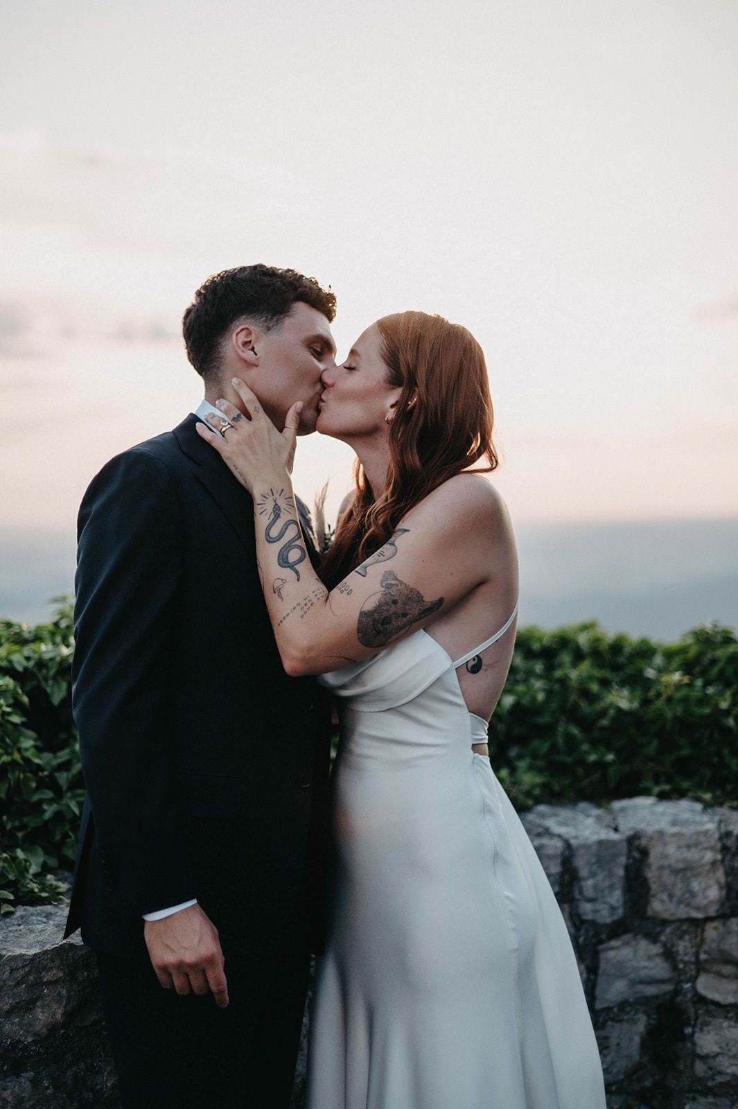 Romantic couple sharing intimate kisses against a stone wall and garden backdrop at sunset.