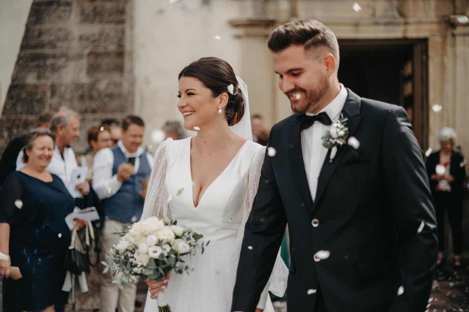Happy newlyweds exit church ceremony with white floral bouquet as guests celebrate in background.