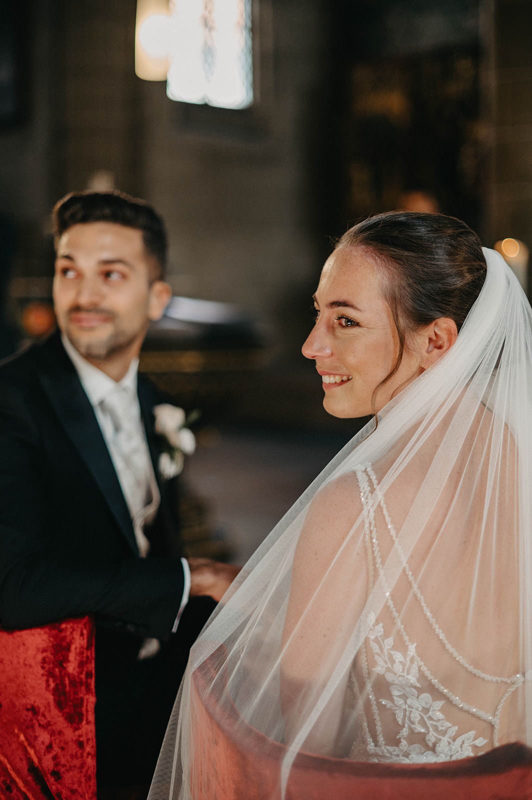 A joyful moment captured during a wedding ceremony with the bride wearing a delicate veil in a dimly lit church setting.