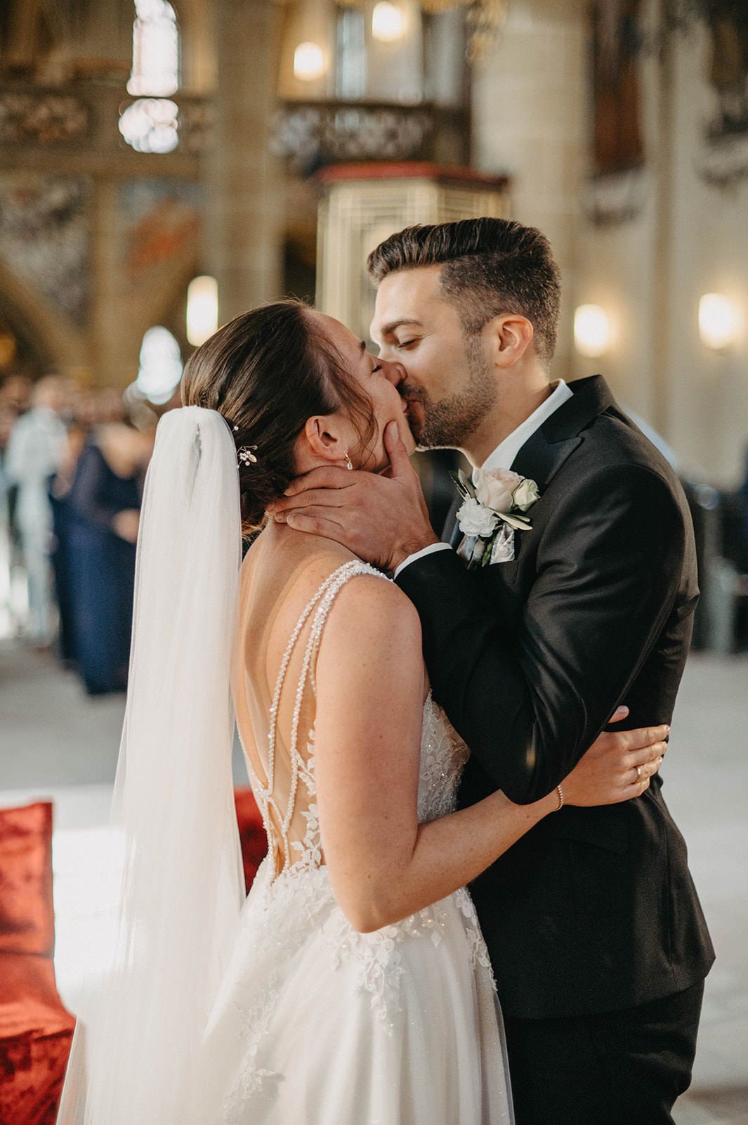 A romantic wedding kiss between newlyweds in a beautifully lit church with elegant decor and guests in the background.