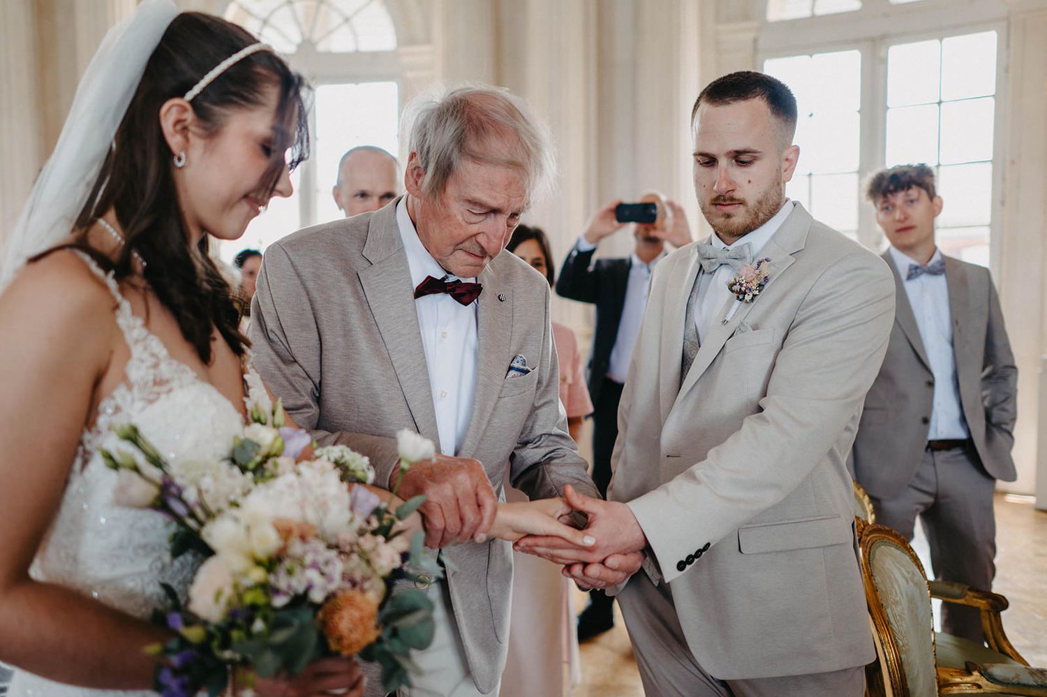 Wedding guests in gray suits gather around a table during a celebration in an elegant venue with natural lighting.