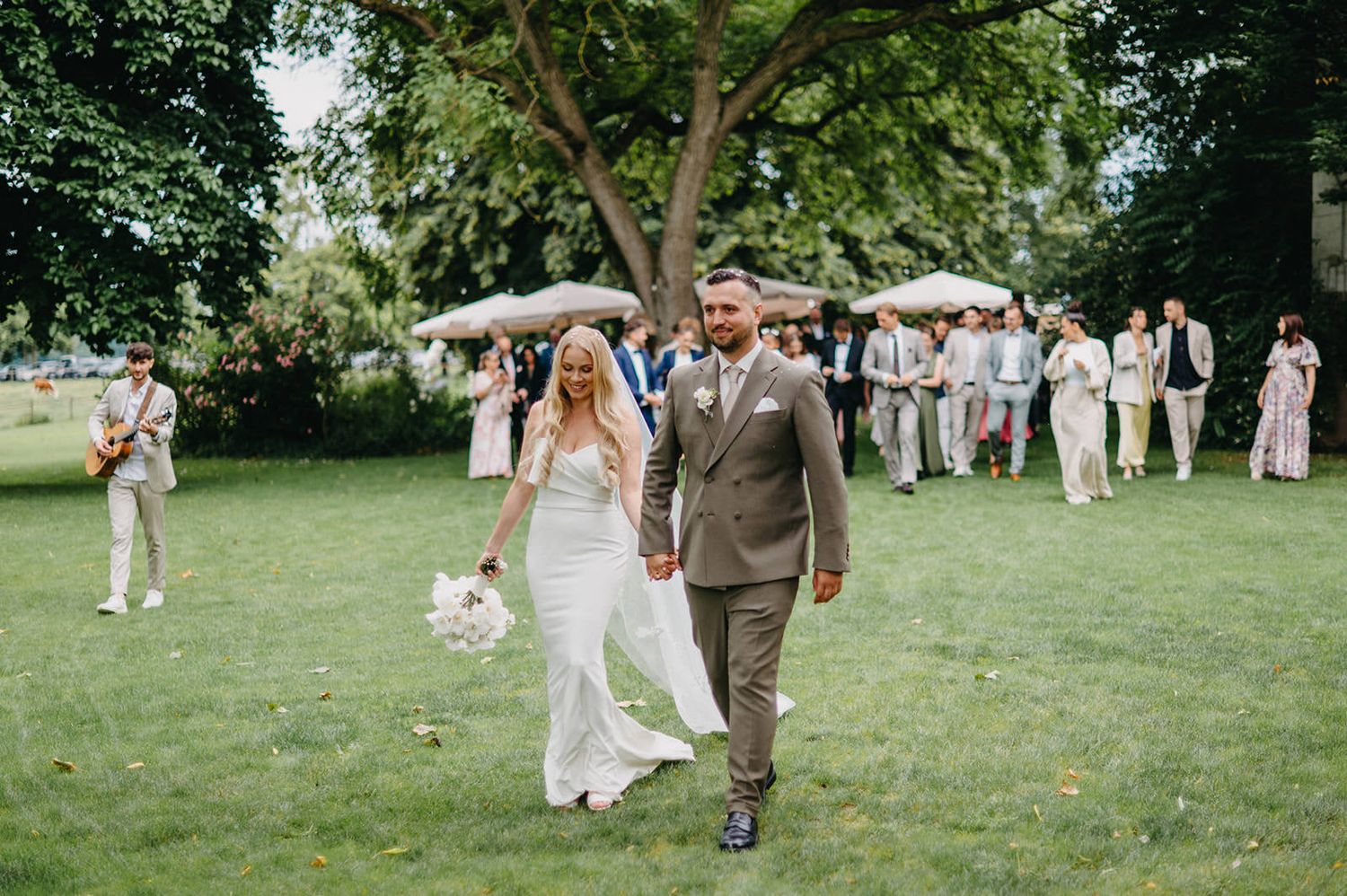 Wedding guests in white attire gather on a lush green lawn during an outdoor summer celebration.