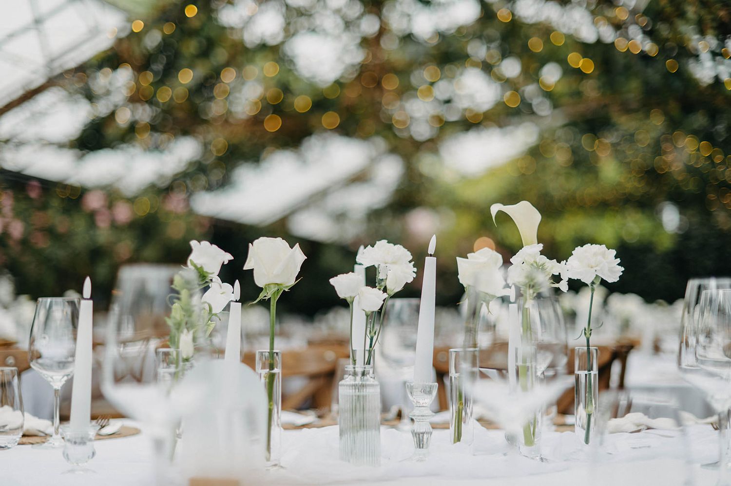 Elegant white floral wedding table decorations with glass vases and candlesticks set against a blurred natural background.