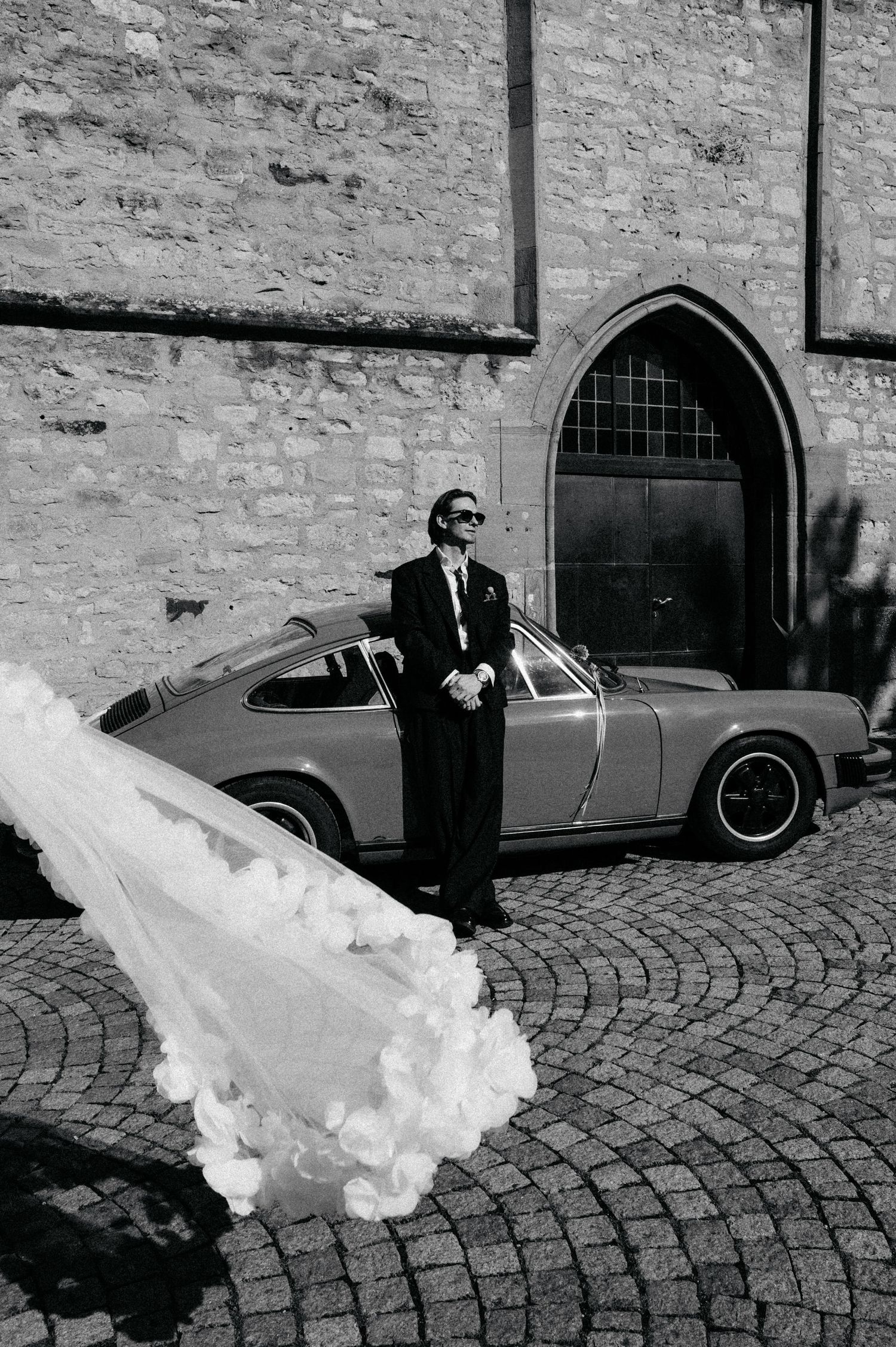 A black and white photo sequence shows smoke billowing from a vintage car parked on cobblestones near a brick wall.