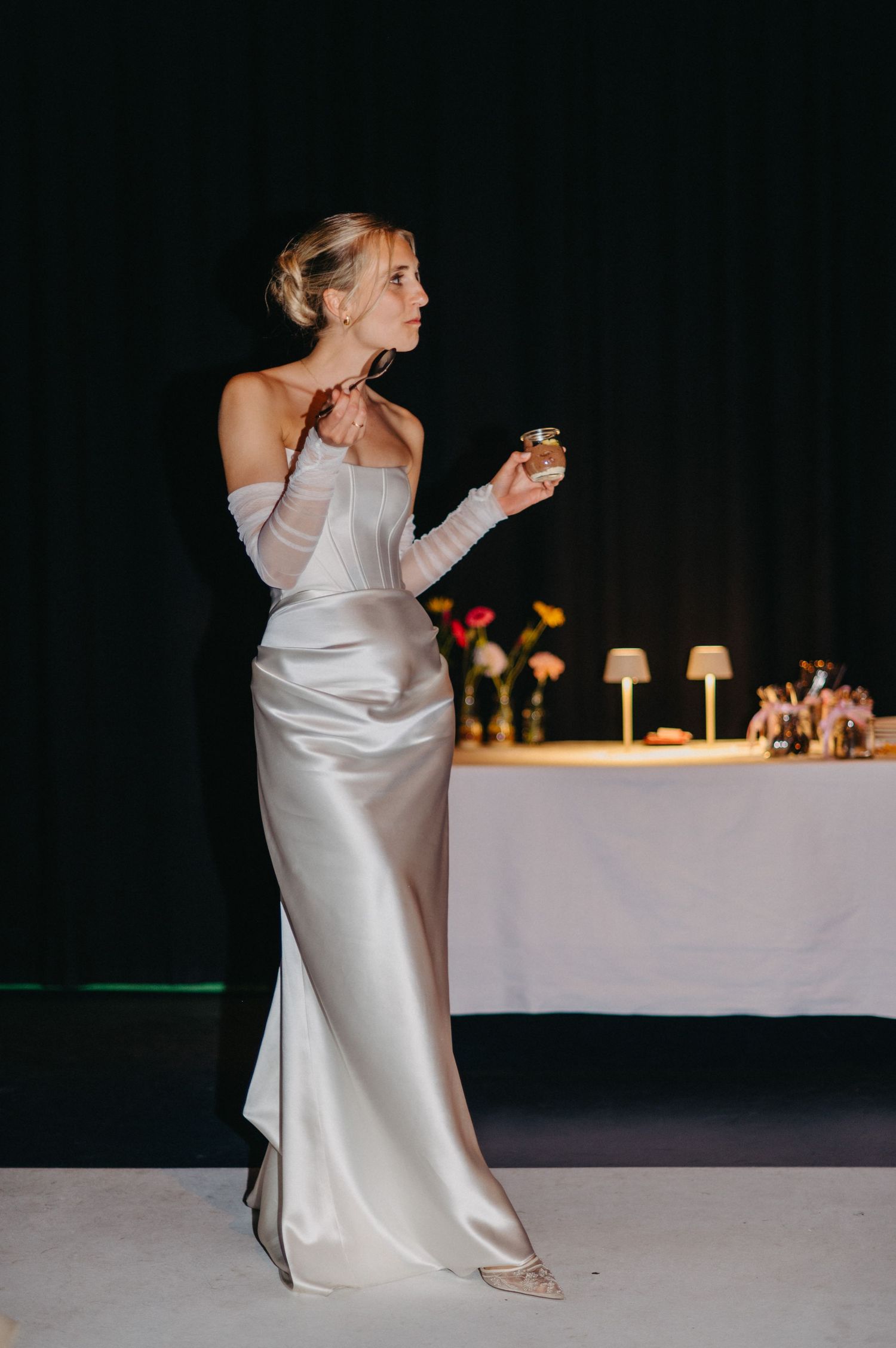 A bride in an elegant white silk gown stands by a decorated table with candles and flowers at an evening reception.