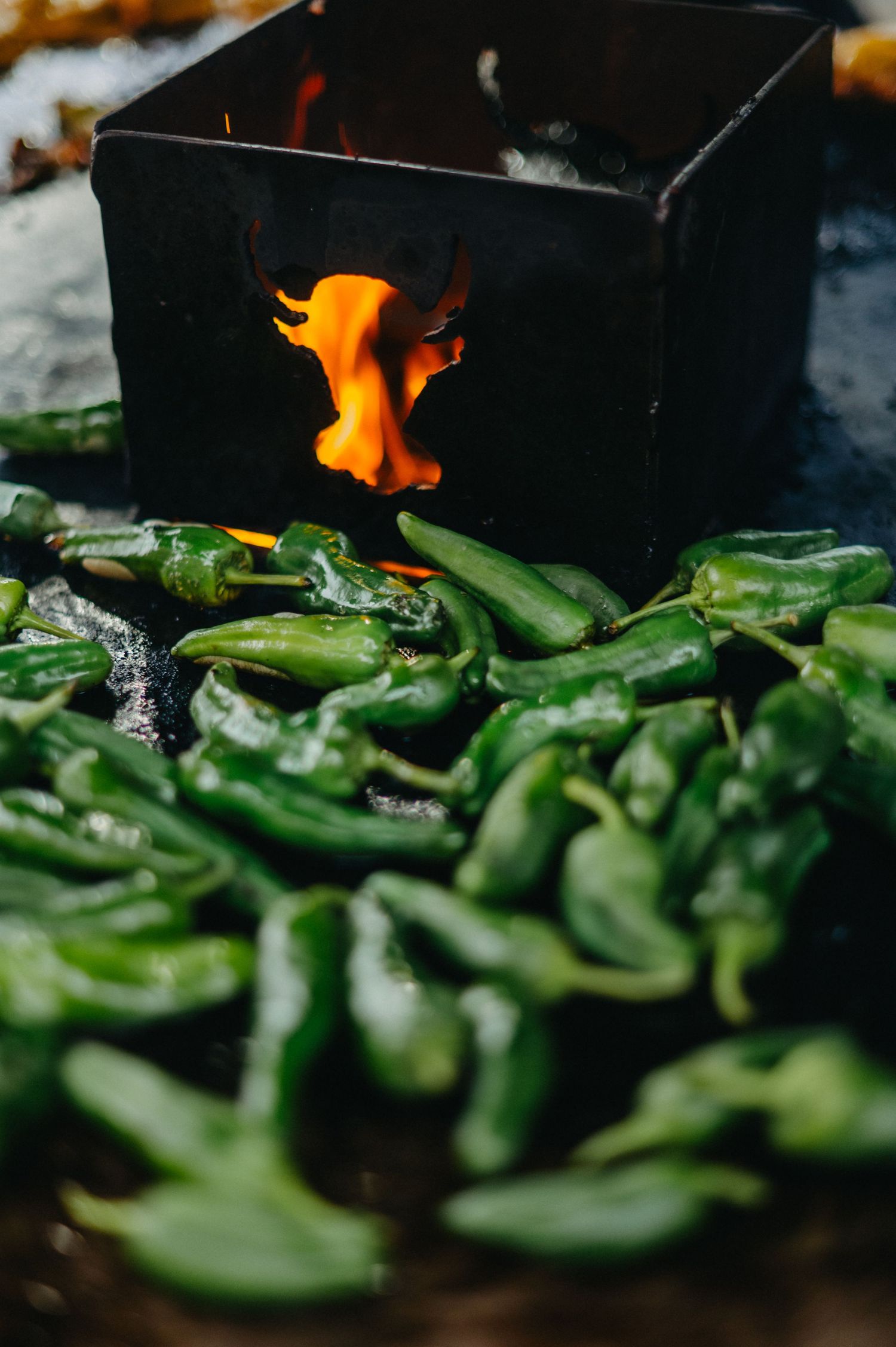Fresh green chili peppers being roasted over an open flame on a dark surface with orange fire visible in the background.
