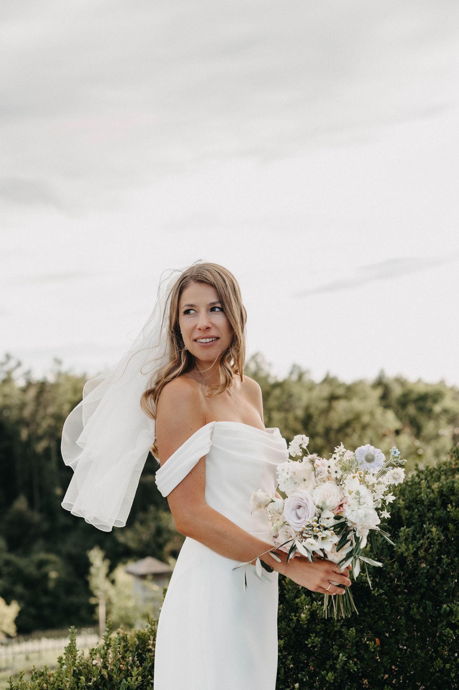Bride holding elegant white floral bouquet against natural outdoor backdrop.