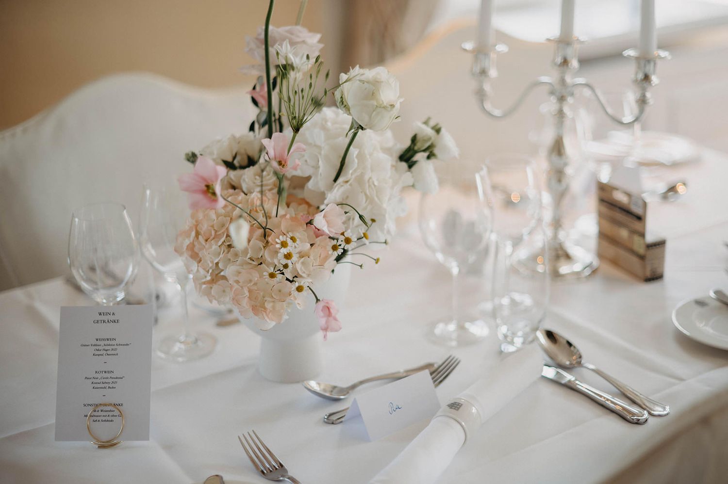 Elegant wedding reception table settings with white floral centerpieces and crystal glassware in a bright venue.