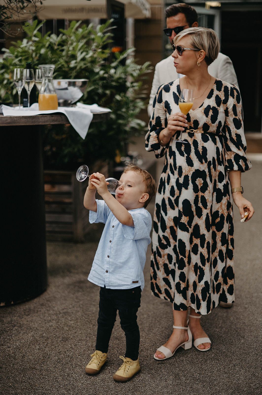 A child in a light blue shirt stands next to someone wearing a leopard print dress while enjoying drinks outdoors.