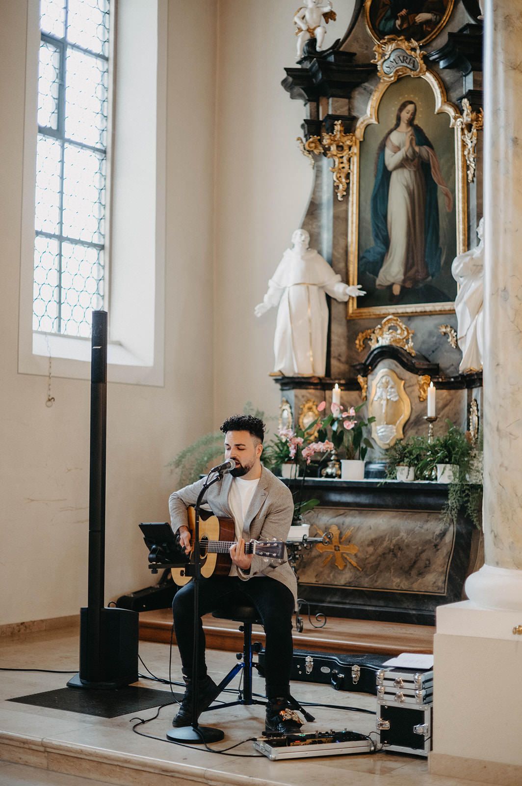 Musician performs with acoustic guitar in ornate church sanctuary with religious statues and stained glass windows.