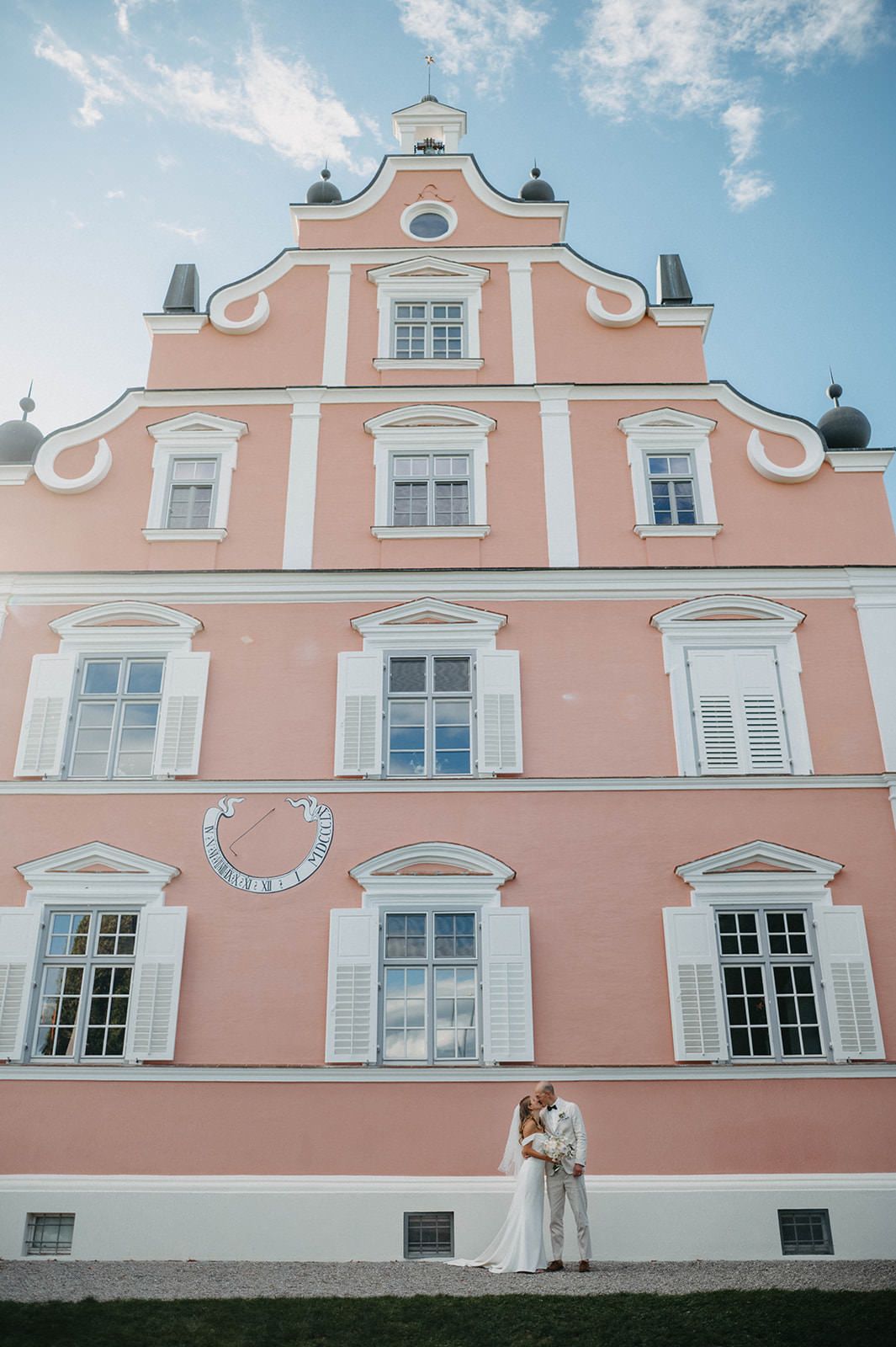A couple stands against a pink historic building with ornate white trim and windows on a sunny day.