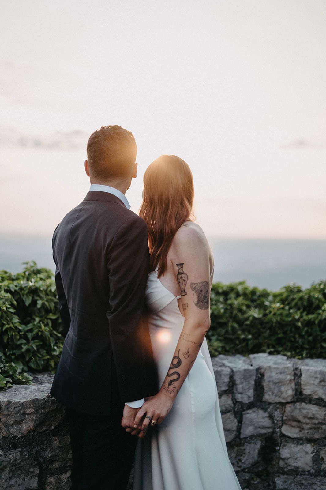 A couple in wedding attire stands together at sunset overlooking a scenic vista, with stone wall and greenery in view.
